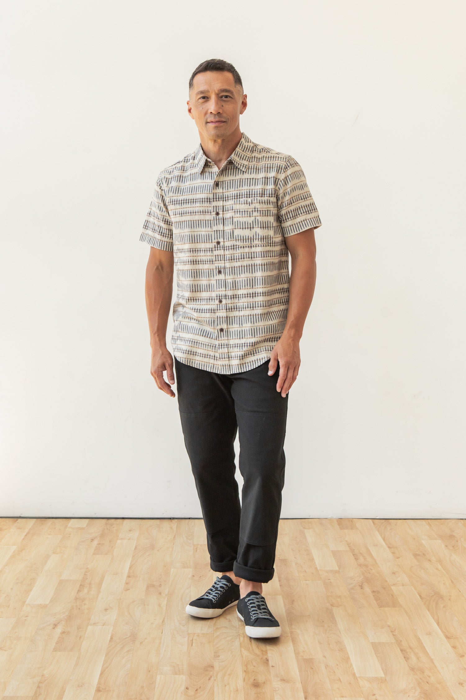 A man stands indoors on a wooden floor, wearing the Bridge & Burn Malcolm Shirt in Mushroom Ikat Stripe, paired with black pants and black sneakers. He faces forward with a neutral expression against a plain white wall.