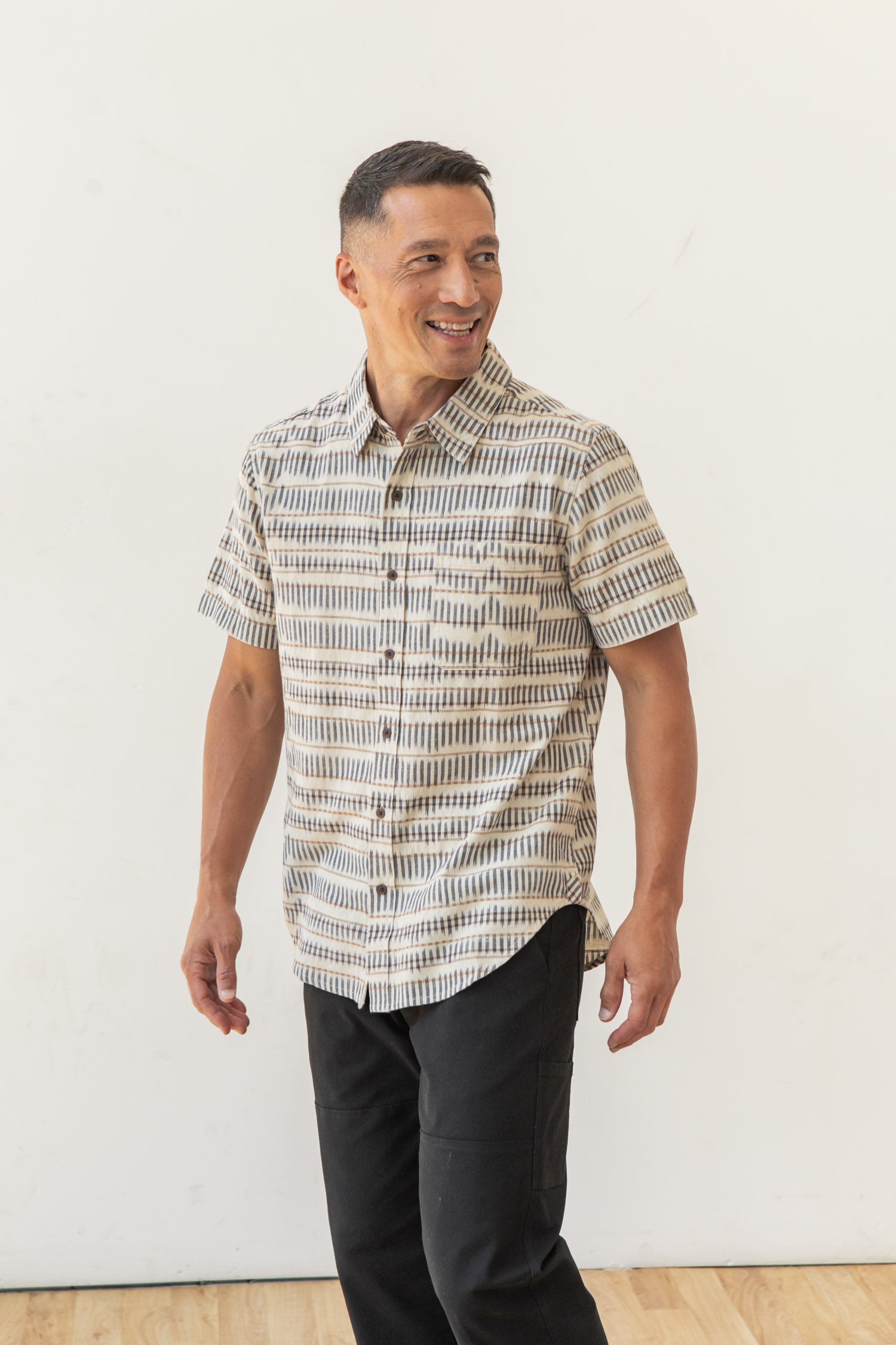 A man stands smiling, wearing the Bridge & Burn Malcolm Shirt in Mushroom Ikat Stripe and black pants, against a plain white background on a wooden floor.