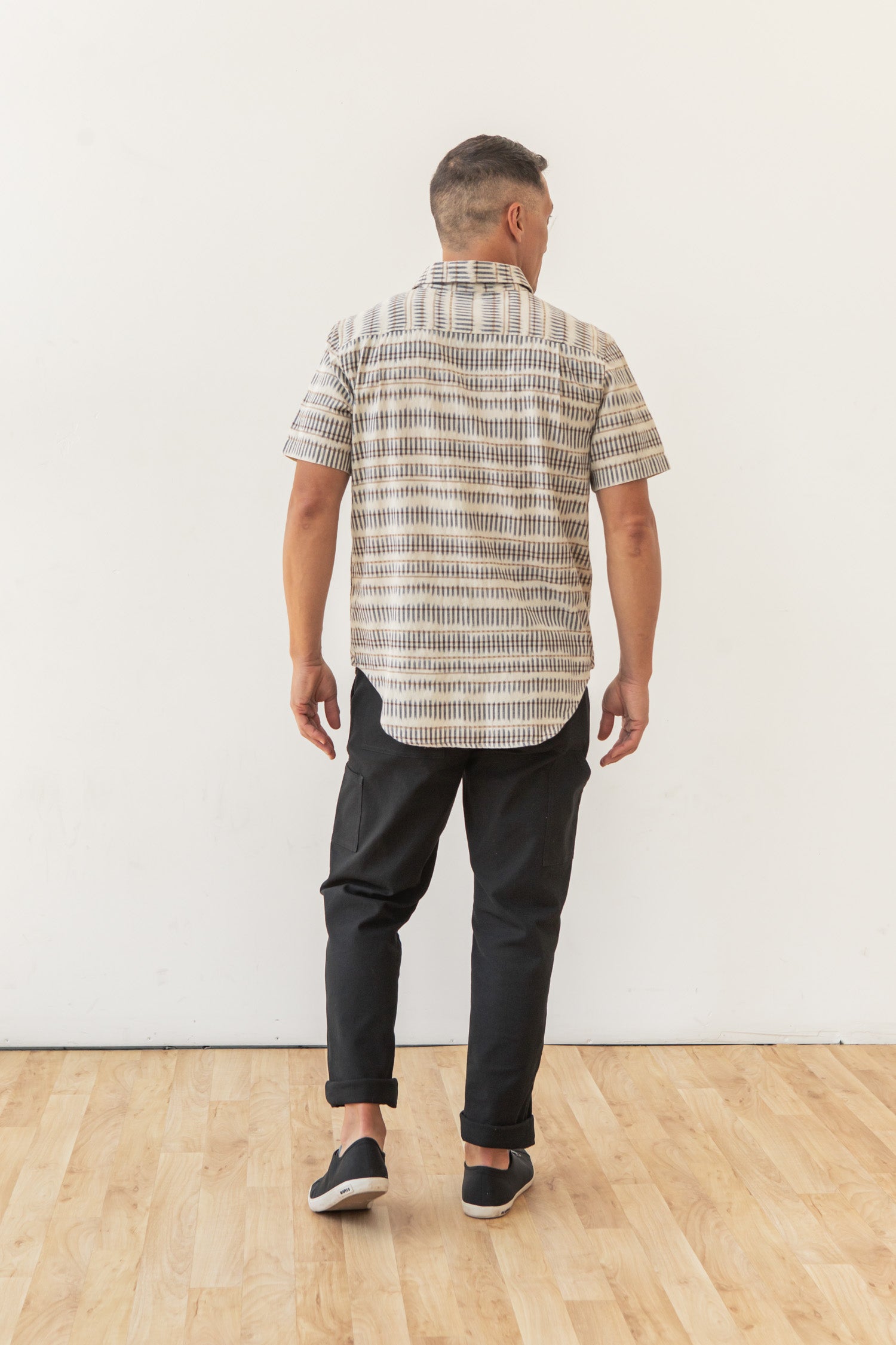 A man stands indoors on a wooden floor, facing away from the camera. He wears the Bridge & Burn Malcolm Shirt in Mushroom Ikat Stripe, paired with black pants and black slip-on shoes, set against a plain white wall.