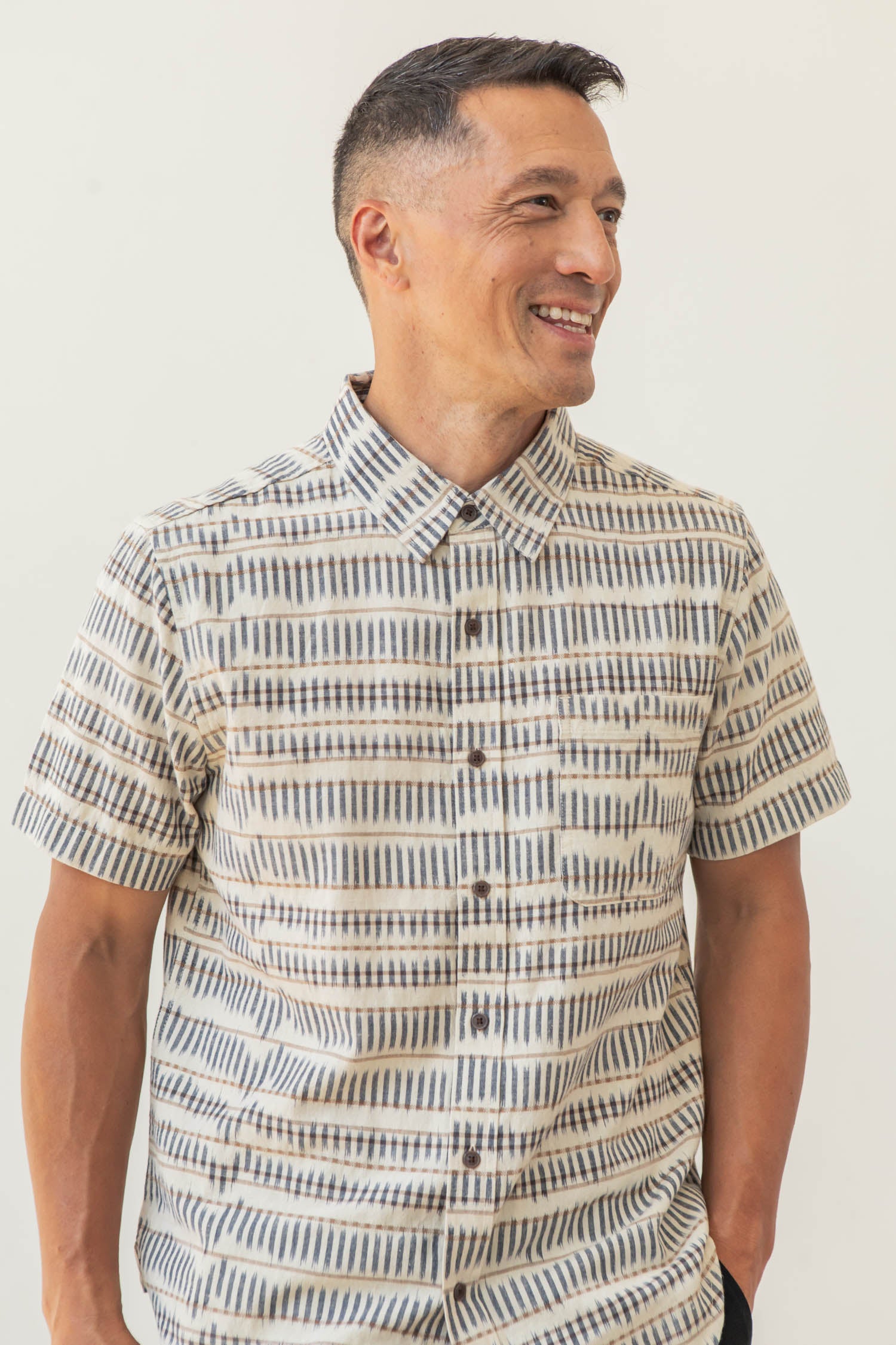 A man stands smiling in the Bridge & Burn Malcolm Shirt in Mushroom Ikat Stripe, a sustainable short sleeve button-up with a neutral geometric stripe pattern, against a plain light background.