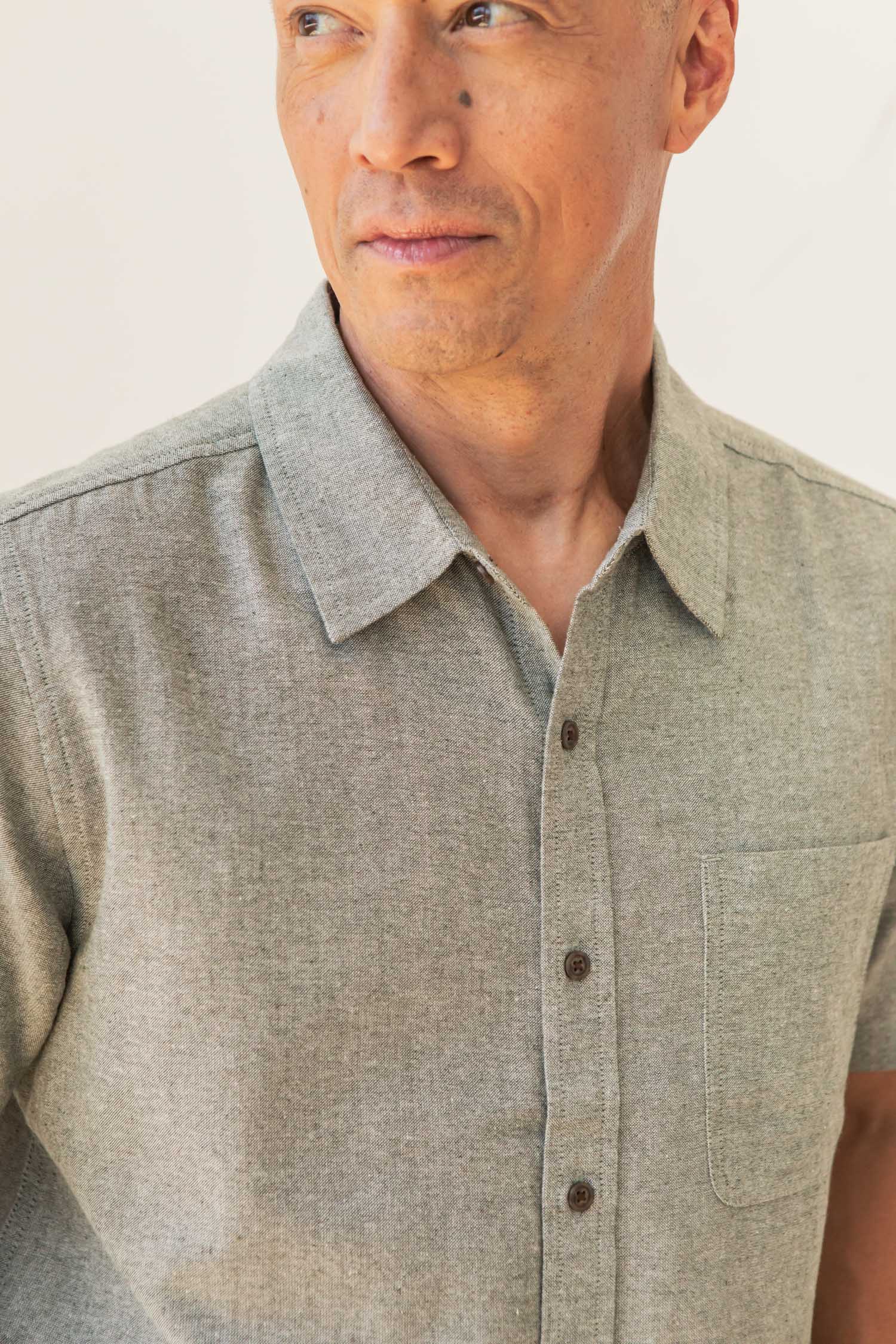 A man wearing the Bridge & Burn Malcolm Shirt in Sage Chambray—a light gray recycled cotton short sleeve button-up with a collar and chest pocket—looks to the side against a neutral background.