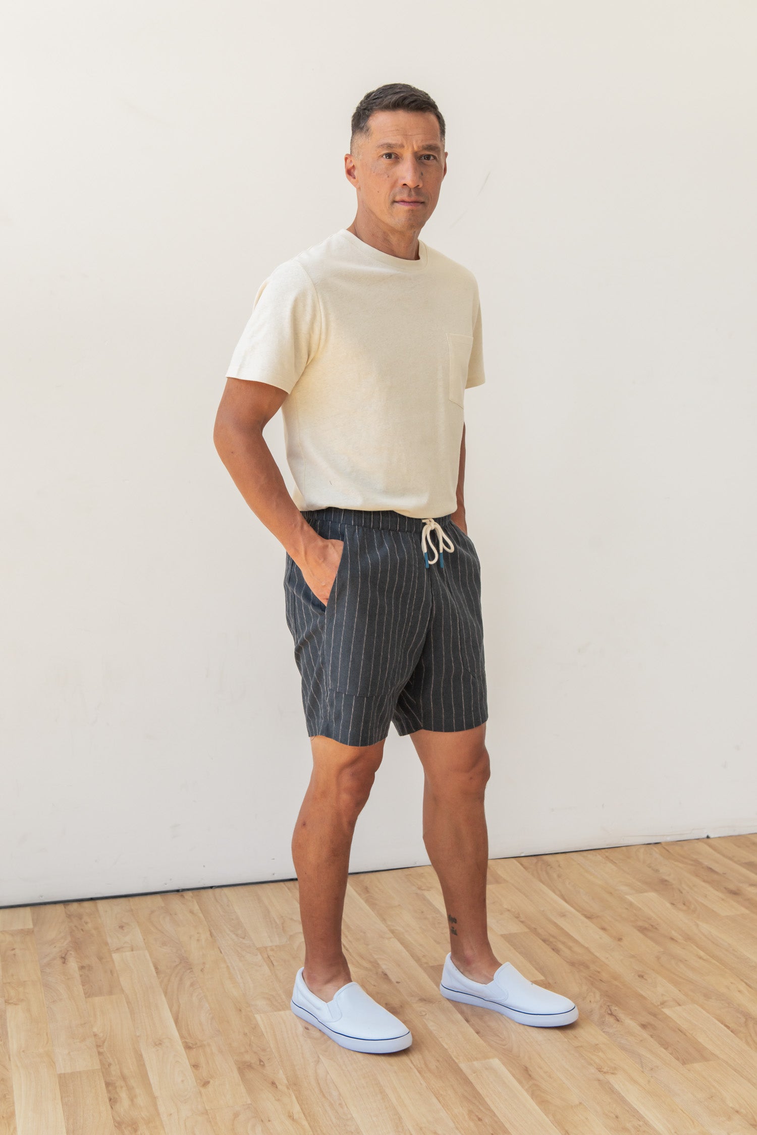 A man stands indoors on a wooden floor against a white wall, wearing the Bridge & Burn Noah Short in Charcoal Stripe with an elastic waist and drawstring, a light beige T-shirt, and white slip-on shoes. His hands are in his pockets.
