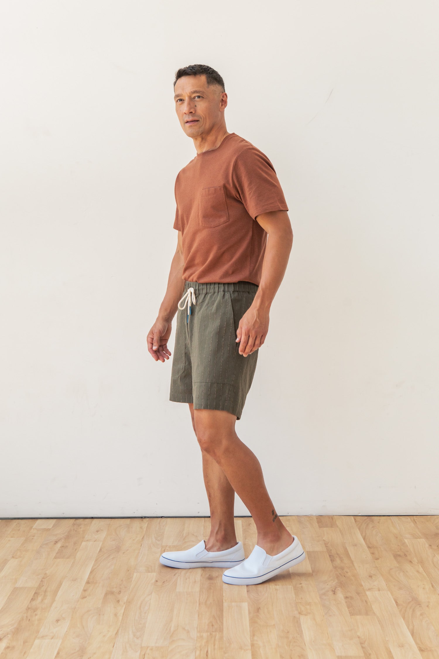 A man stands indoors on a wooden floor against a plain white wall, wearing Bridge & Burn's Noah Short in Olive Stripe with an elastic waist, a rust t-shirt, and white slip-on shoes. He faces slightly to the side and looks at the camera.