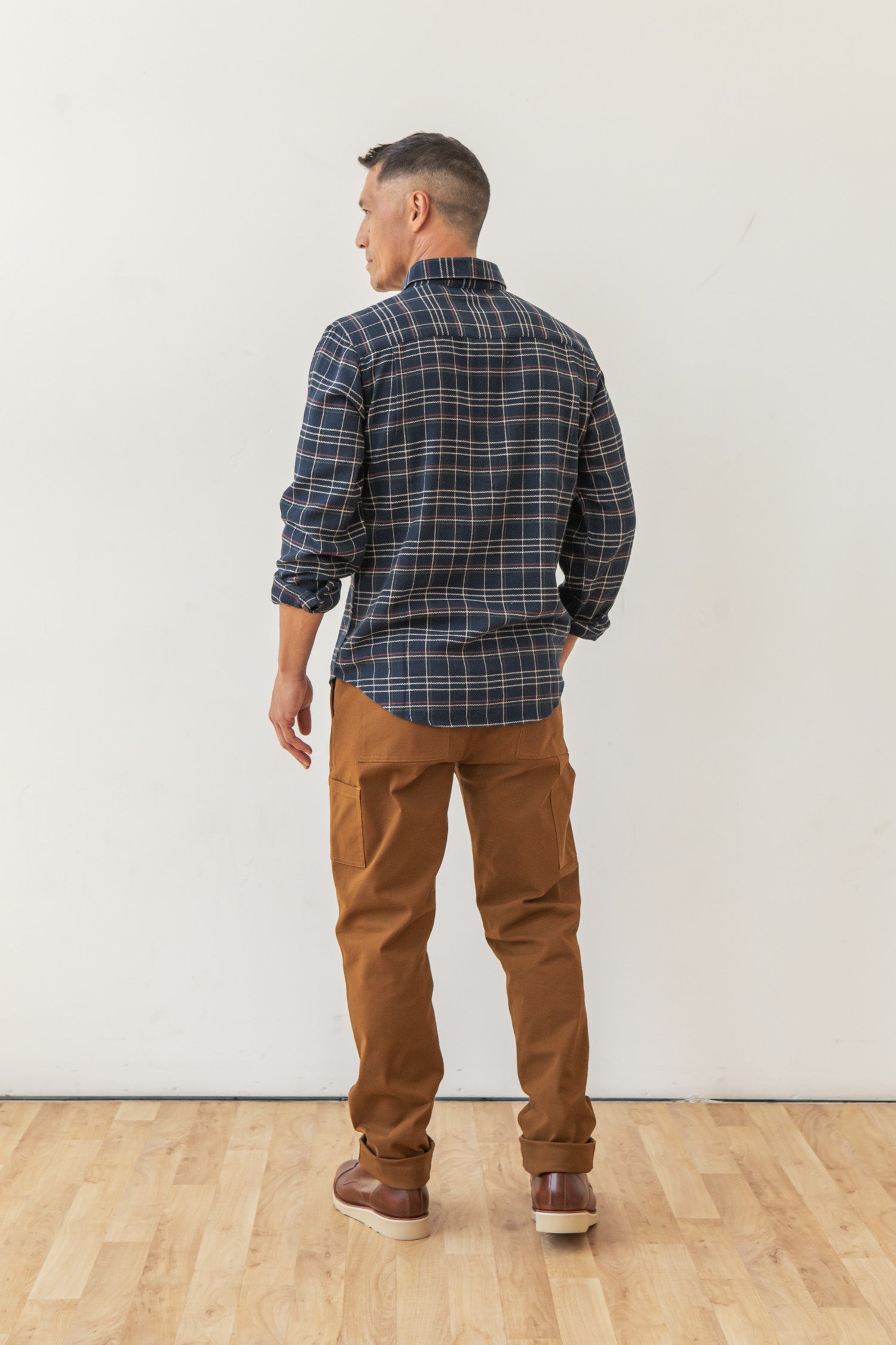 A man stands on a wooden floor facing away, wearing the Bridge & Burn Sutton Slim Shirt in Dusk Herringbone Plaid, along with brown pants and shoes. The backdrop is a plain white wall, creating a clean, minimalist look.