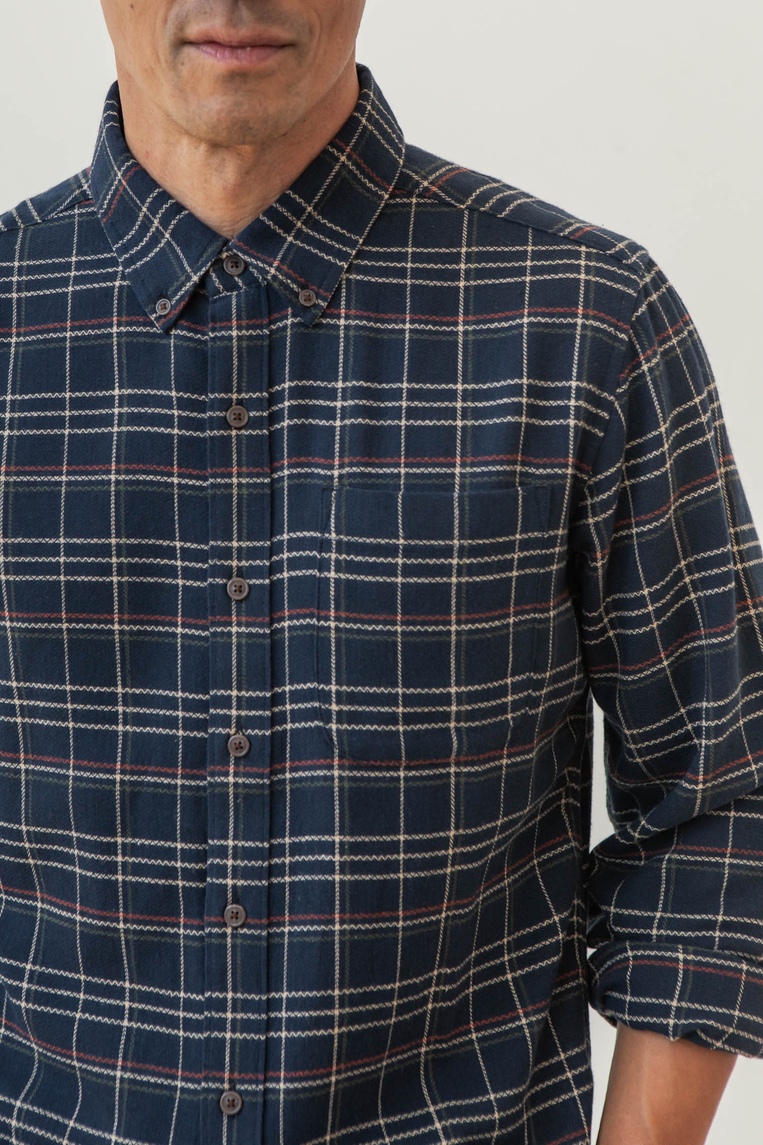 A person wears the Bridge & Burn Sutton Slim Shirt in Dusk Herringbone Plaid, a navy slim-fit button-up with red, white, and beige lines. The image shows their upper body from chest to just above the mouth, with one sleeve slightly rolled up.