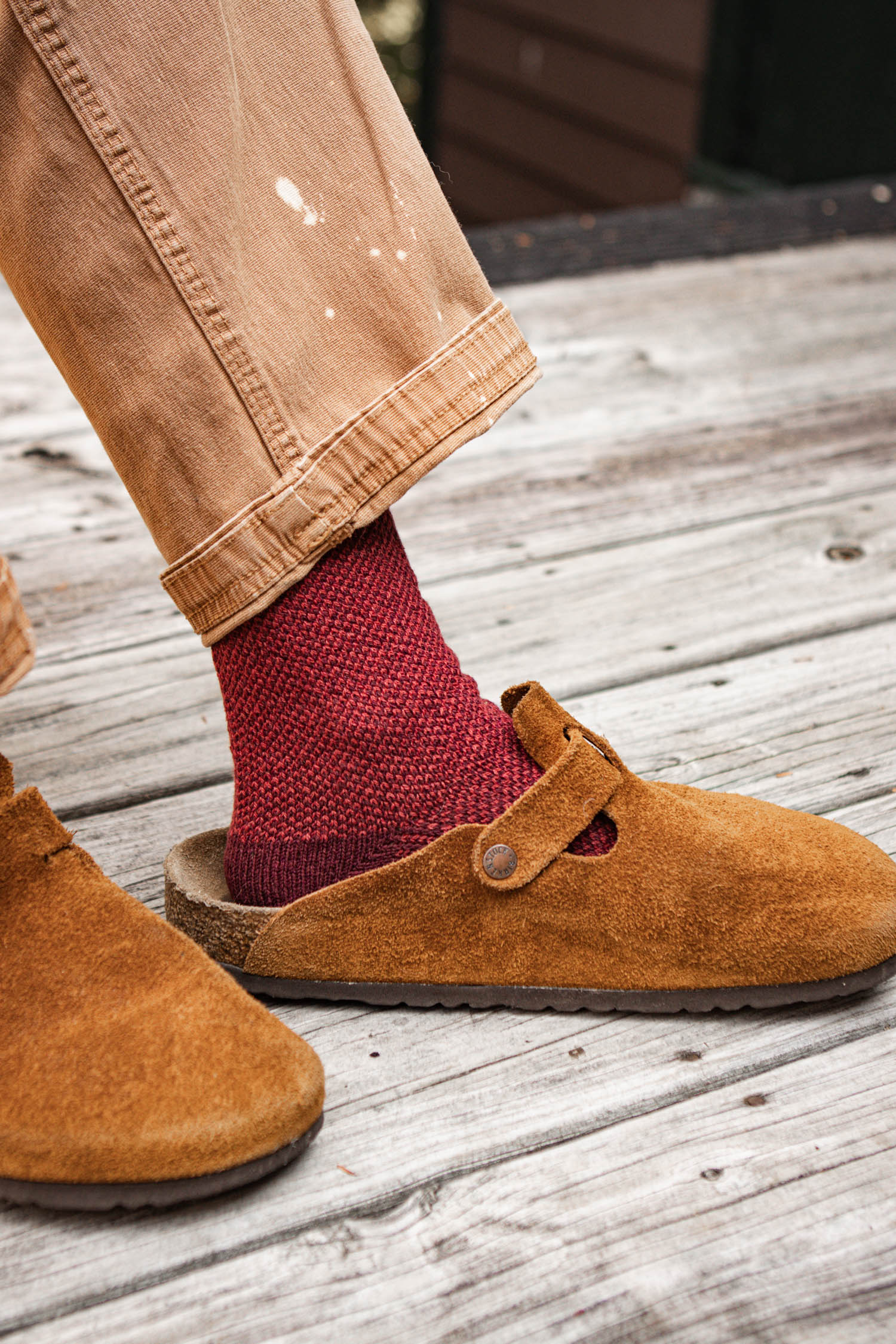 A person wearing light brown pants, Bridge & Burn Helix Sock in Burgundy, and tan suede clogs stands on a weathered wooden deck.