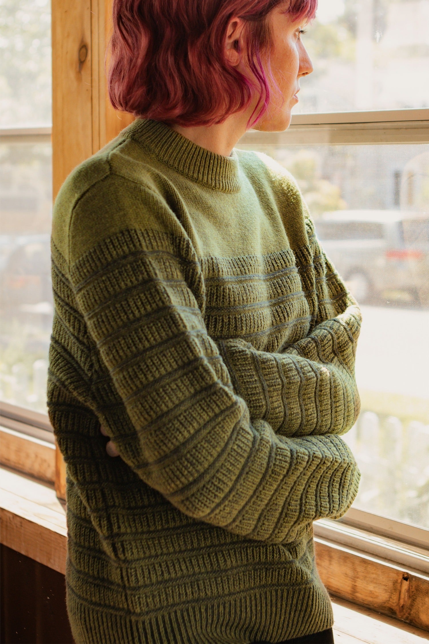 A person with short, wavy, pink hair stands indoors by a sunlit window, arms folded, wearing the Bridge & Burn Sabine Sweater in Olive.
