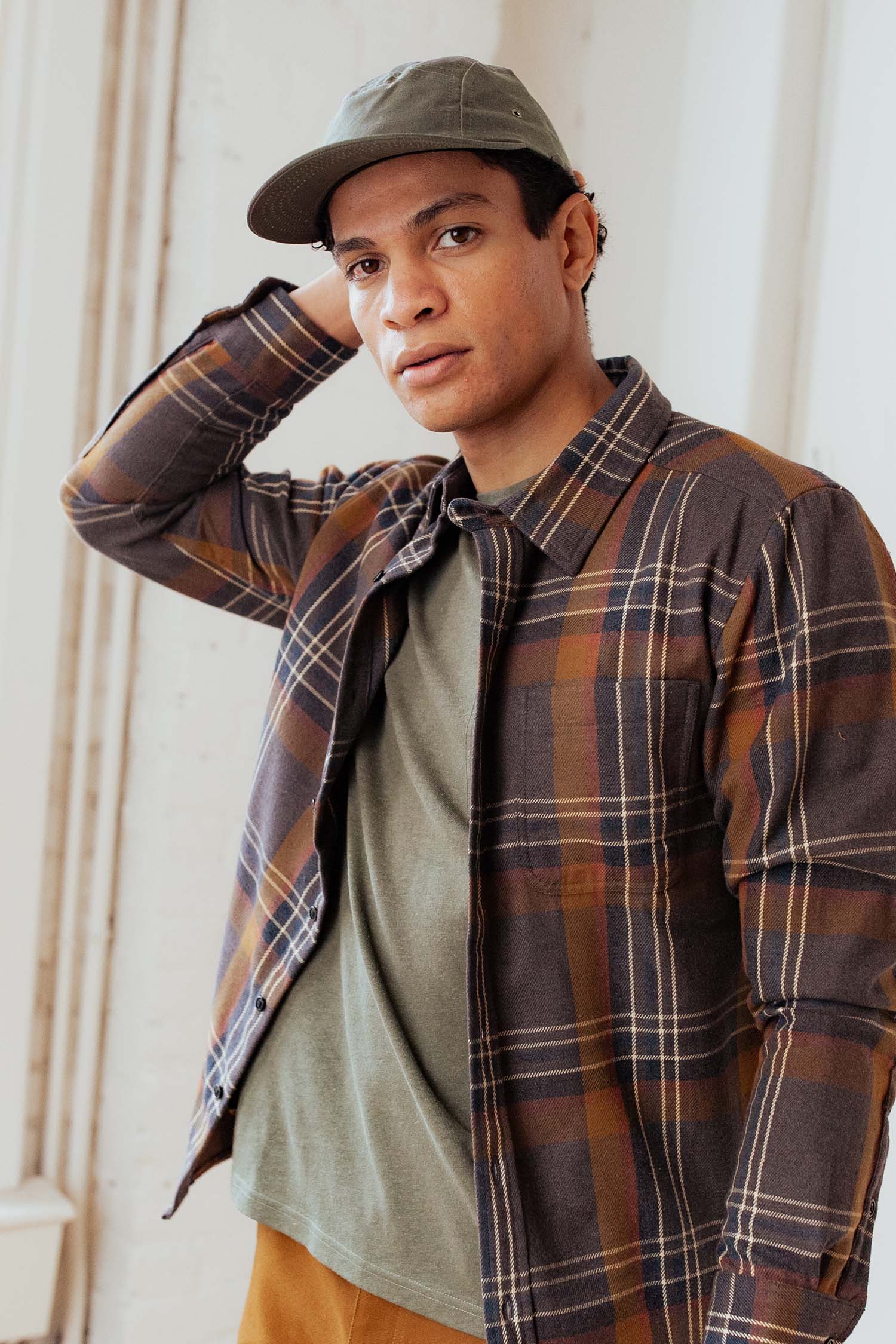 A person wearing the Bridge & Burn Camper Waxed 5 Panel Cap in Moss, an olive green shirt, and a brown plaid jacket stands indoors with one hand behind their head, looking at the camera.