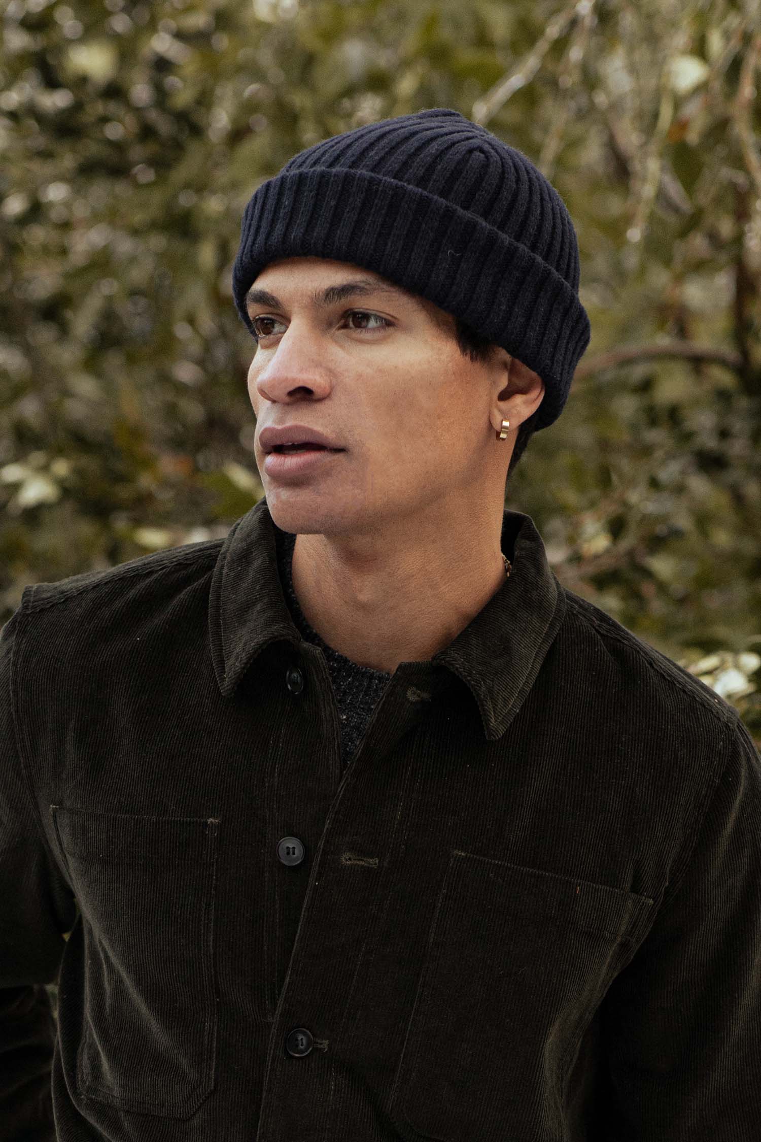 A person wearing the Bridge & Burn Chunky Ribbed Beanie in Dark Navy stands outdoors before blurred greenery, dressed in a dark corduroy jacket and an earring, gazing slightly sideways with a neutral expression.