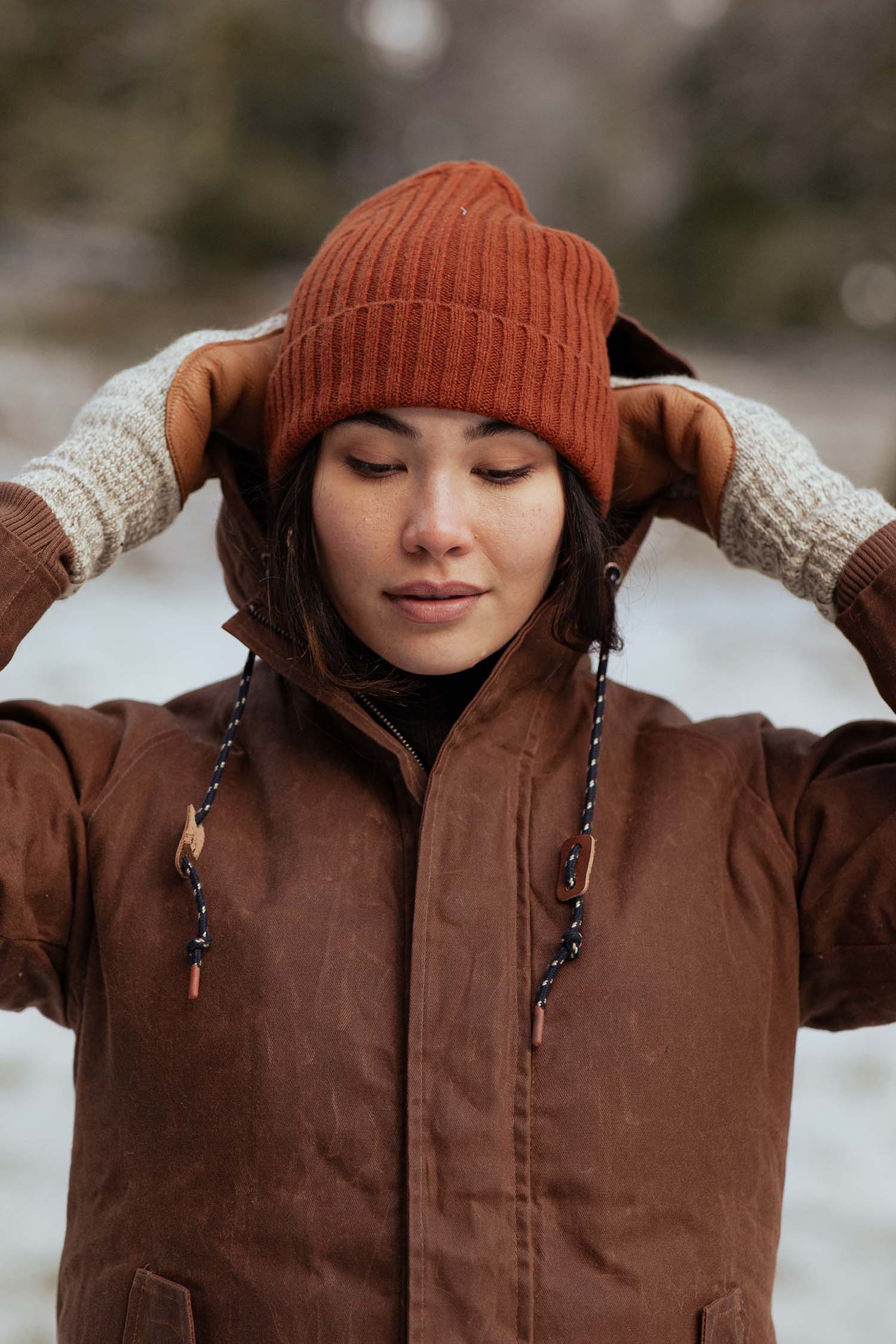 A person in a brown winter coat and beige gloves stands in the snow, gently holding the sides of their Bridge & Burn Chunky Ribbed Beanie in Sienna.