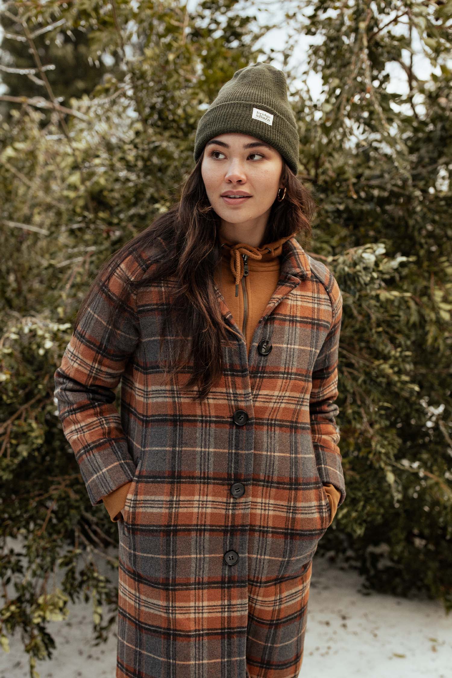 Wearing a Bridge & Burn Wool Watch Cap in Olive Drab, a person stands outside before snow-dusted greenery, hands tucked in their long plaid coat’s pockets, brown hoodie underneath, gazing thoughtfully to the side.