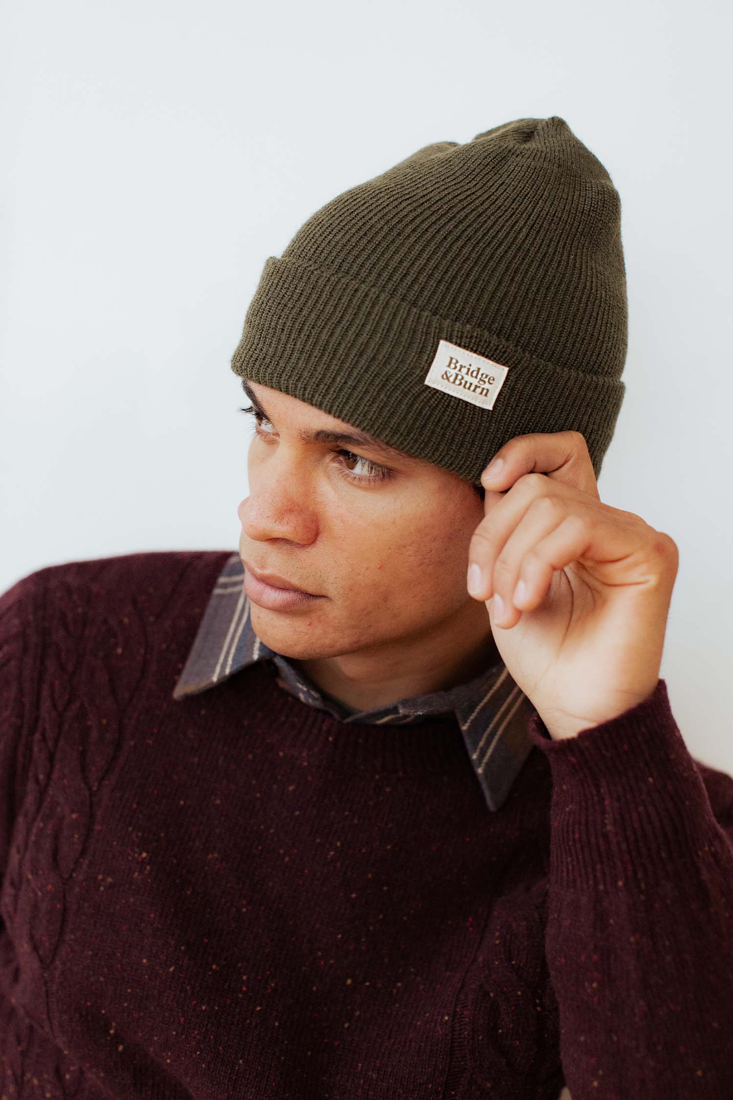 A person in a Bridge & Burn Olive Drab Wool Watch Cap and a maroon cable-knit sweater looks to the side while adjusting the beanie.