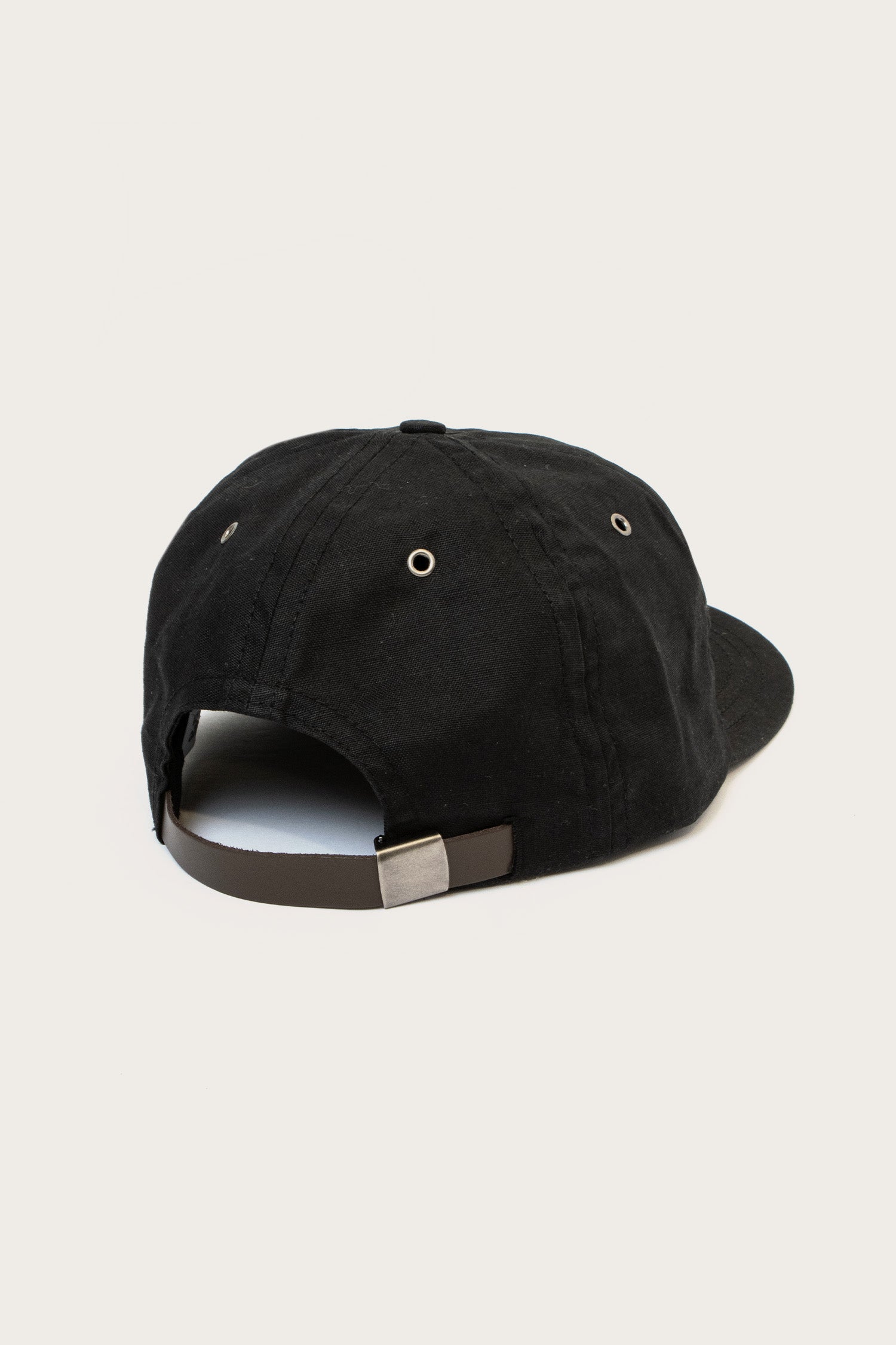 The Bridge & Burn B Flat Waxed Cap in Black features a metal buckle and adjustable strap at the back, metal eyelets for ventilation, and a curved brim—shown here against a simple light background.