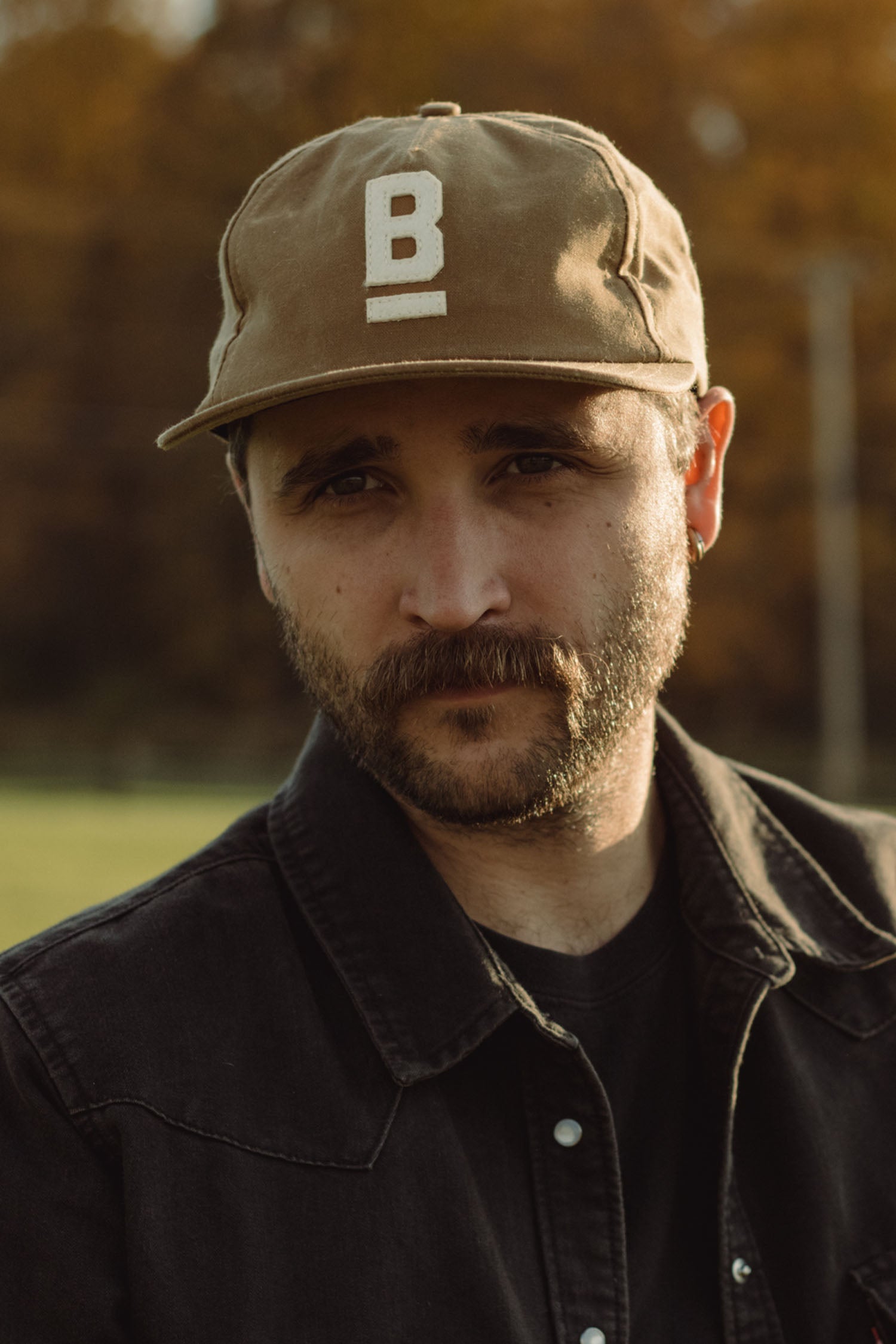 A man with a mustache and beard wears the Bridge & Burn B Flat Waxed Cap in brown—a classic British Millerain canvas hat with a white "B"—and a black jacket, standing outdoors with blurred autumn trees in the background.
