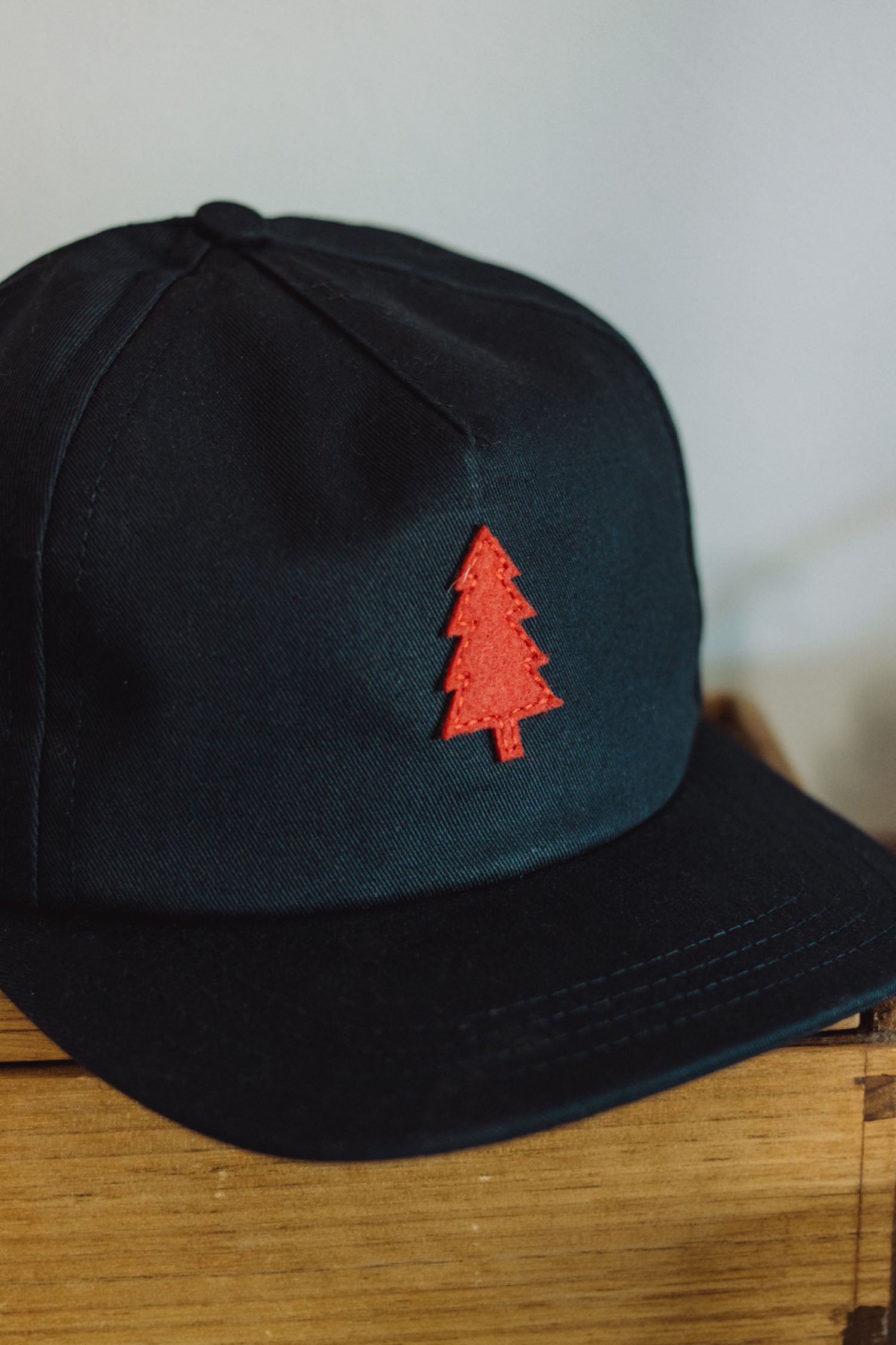 A navy cotton twill Tree Cap by Bridge & Burn, featuring a red embroidered pine tree emblem and a leather adjustment strap, is displayed on a wooden surface.