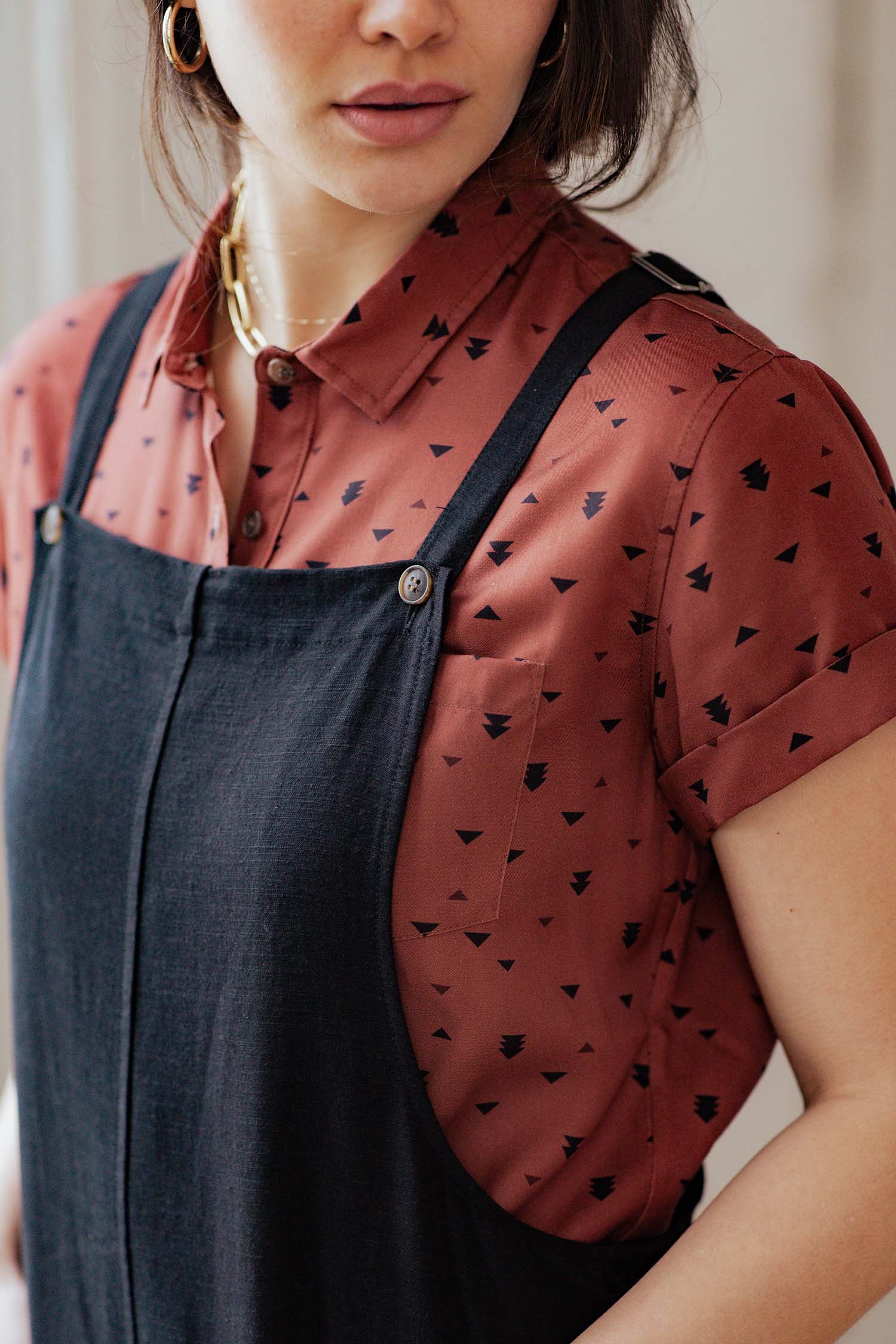 A woman with short hair wears the Bridge & Burn Edie Overall in black over a rust short-sleeve shirt with a black geometric pattern. She accessorizes with gold hoop earrings and a gold necklace; only her upper torso and face are visible.