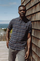 A man in the Bridge & Burn Jordan Slim Shirt in Dusk Herringbone Plaid and gray pants leans against a wooden shingle wall on a sunny deck, with glimpses of the ocean and a greenhouse in the background.