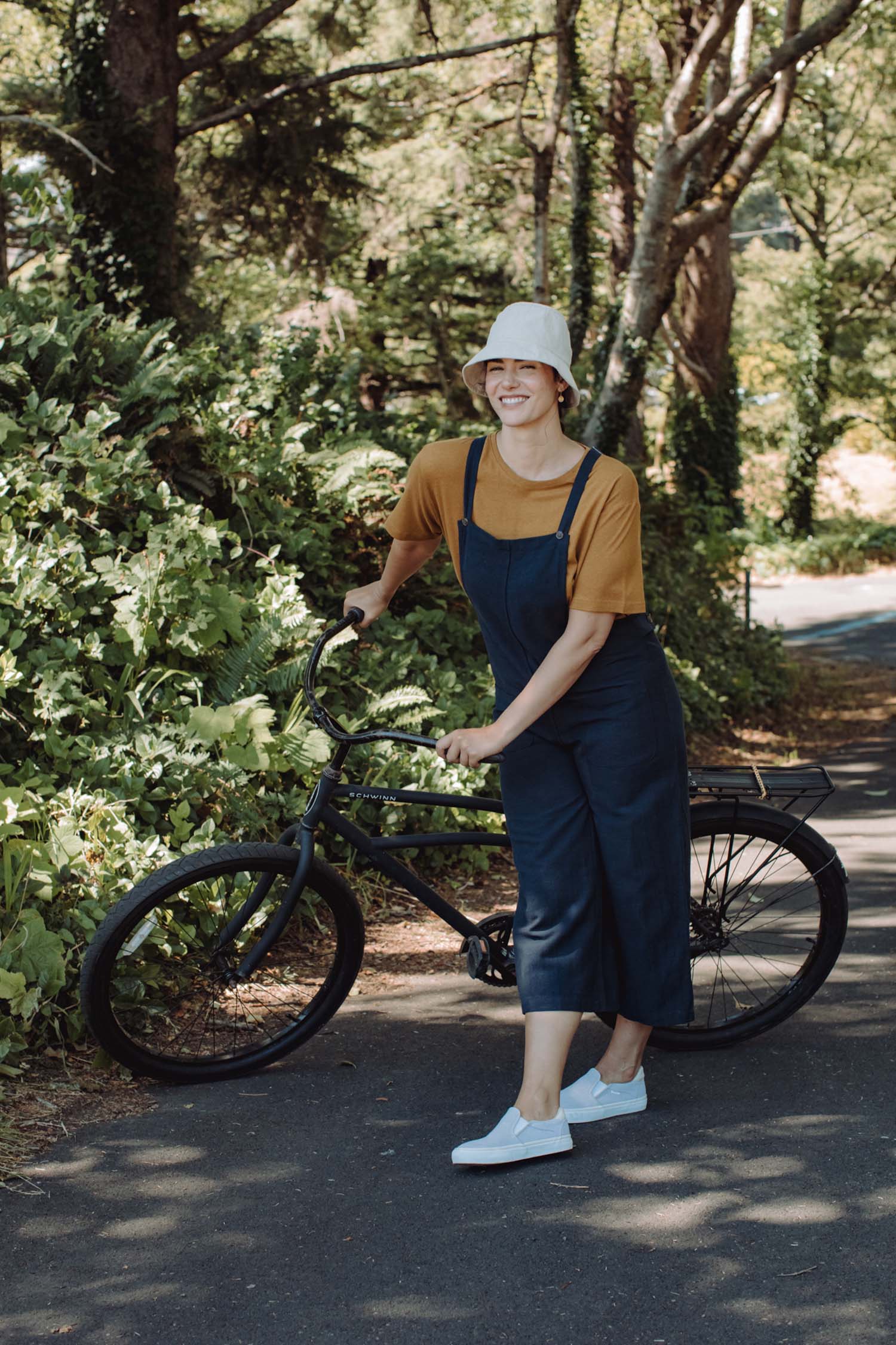 Wearing a bucket hat, orange shirt, and Bridge & Burn's Edie Overall in Navy, a person stands smiling on a sunny paved path with a black bicycle, surrounded by lush green trees and foliage.