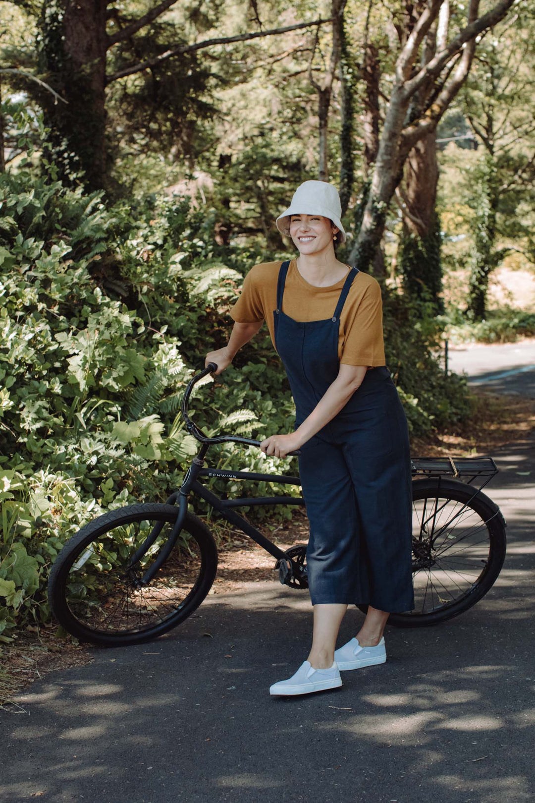 Wearing a bucket hat, orange shirt, and Bridge & Burn's Edie Overall in Navy, a person stands smiling on a sunny paved path with a black bicycle, surrounded by lush green trees and foliage.