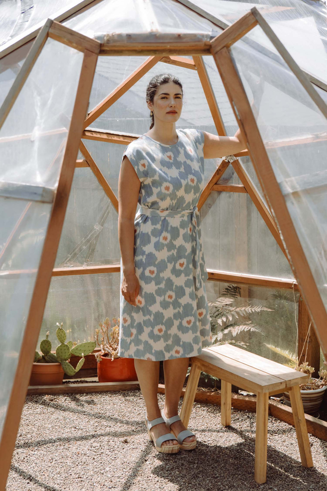A woman wears the Bridge & Burn Eloise Dress in Polar Blossom inside a small wooden greenhouse, resting her hand on the frame by a bench, with potted plants and cacti in the background.