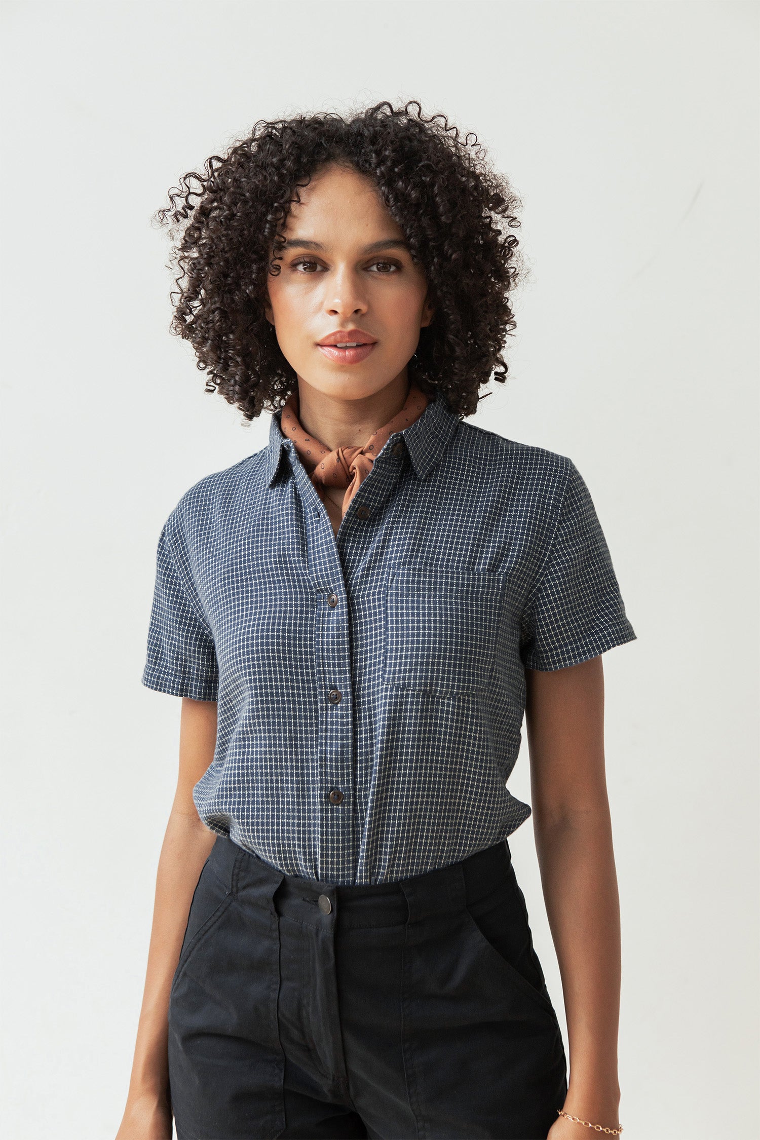 A woman with curly hair wears the Bridge & Burn Lana Shirt in Navy Grid, tucked into dark pants with a light brown neck scarf, standing against a plain light background.