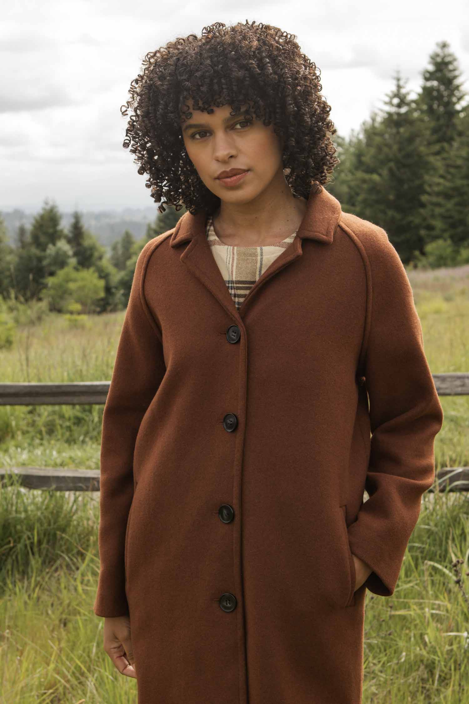 A person with curly hair wears the Bridge & Burn Alberta Car Coat in Fired Brick over a plaid shirt, standing outdoors by a wooden fence with trees and greenery in the background.