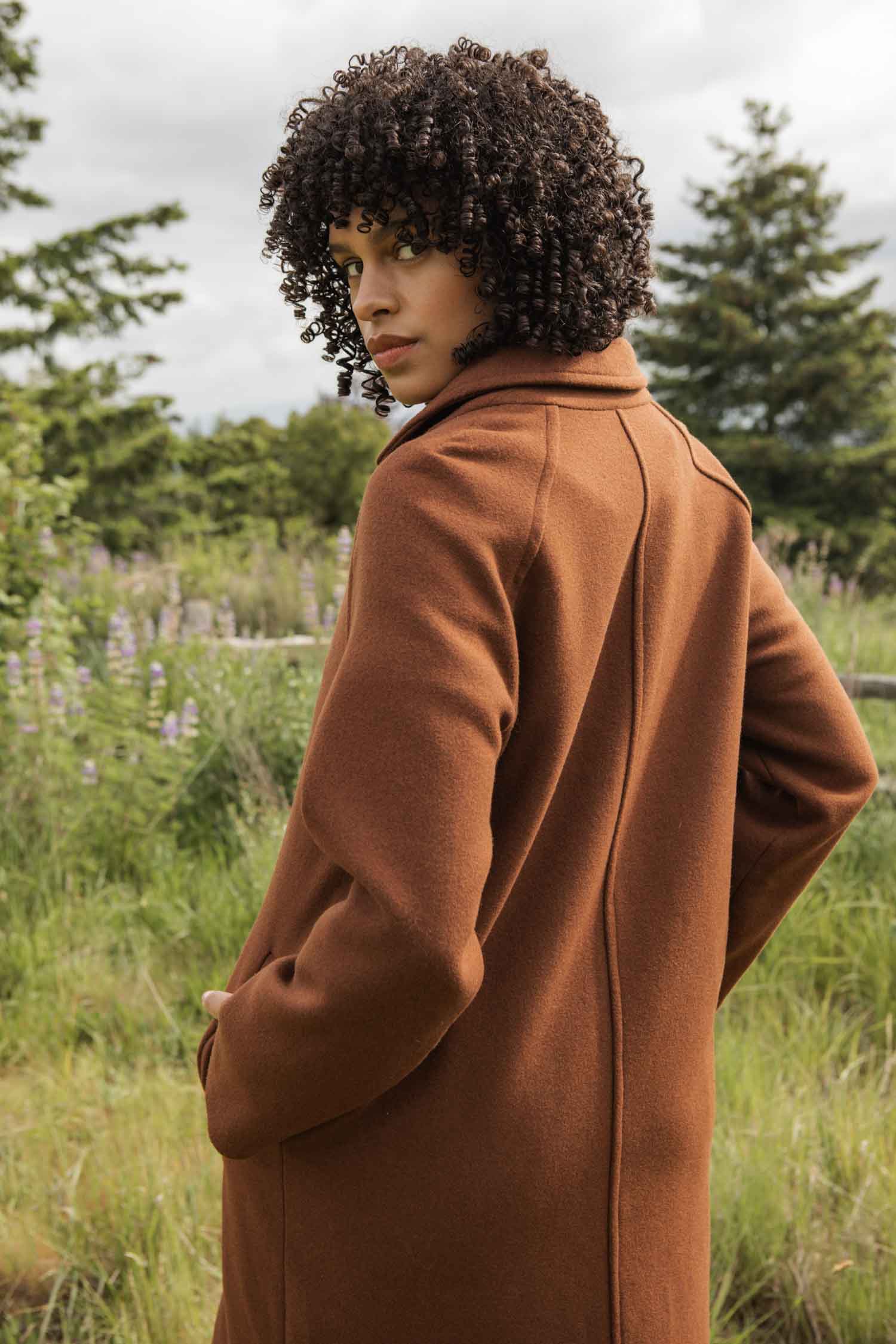 Wearing the Bridge & Burn Alberta Car Coat in Fired Brick, a person with curly hair stands outdoors among grass, trees, and wildflowers, looking over their shoulder at the camera.