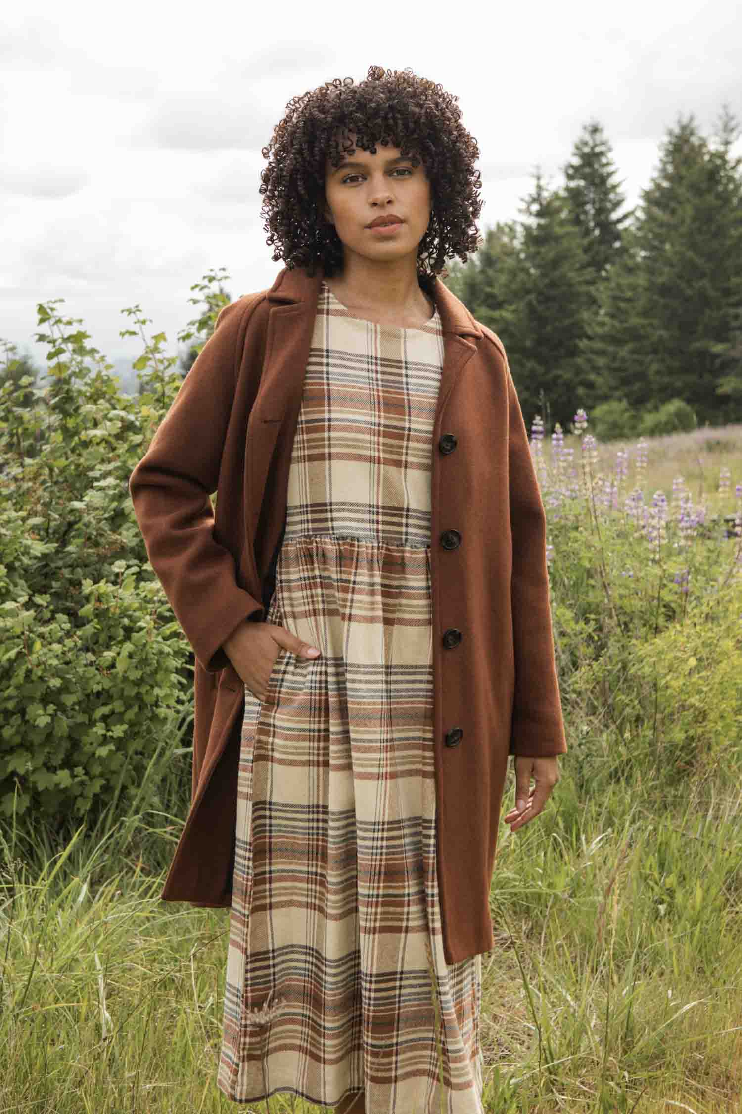 A woman with curly hair stands in a grassy field under a cloudy sky, wearing the Bridge & Burn Alberta Car Coat in Fired Brick over a beige and brown plaid dress. Green trees and plants are visible in the background.
