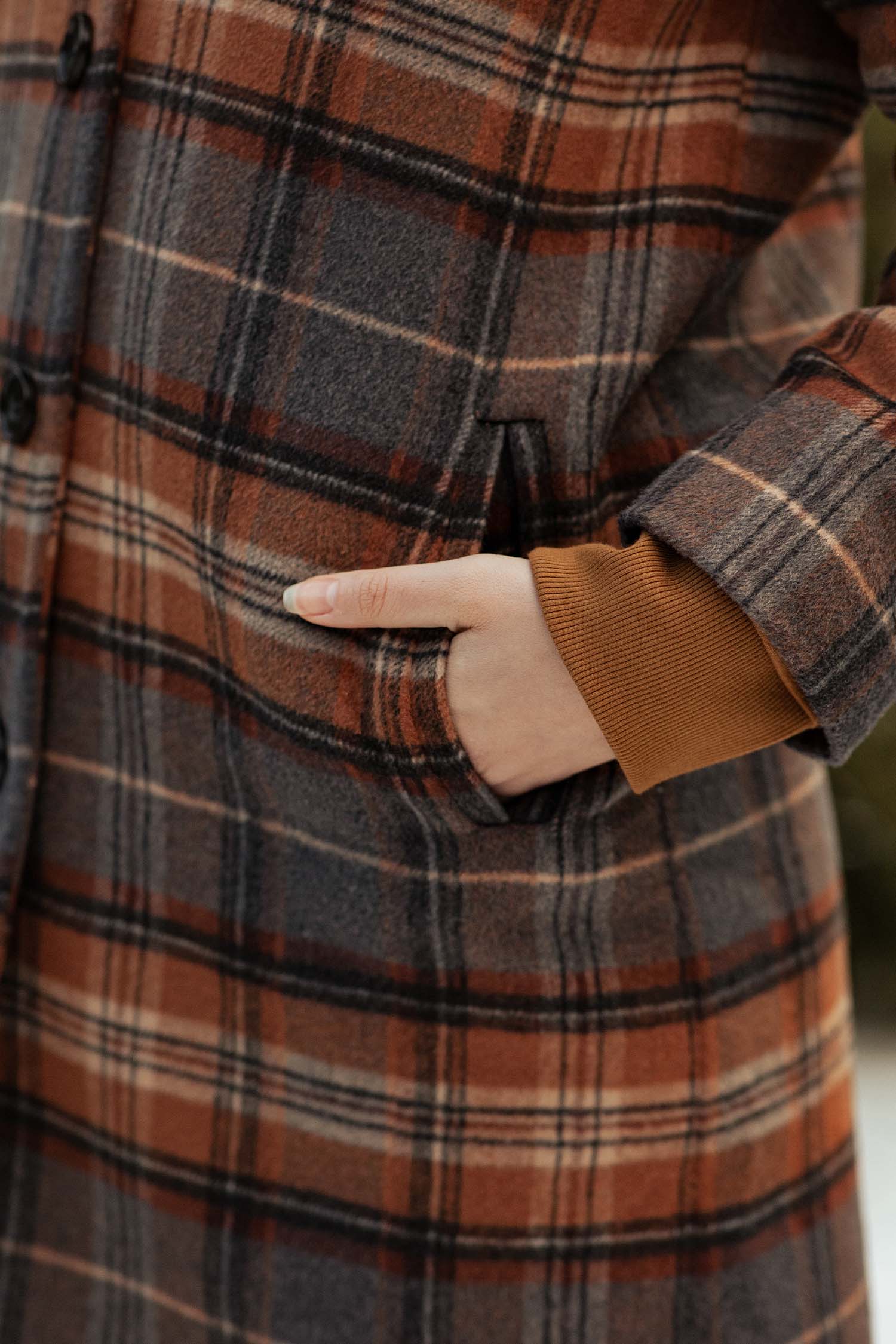A close-up shows a hand in the pocket of Bridge & Burn’s Alberta Car Coat in Dorset Brushed Plaid—heavyweight wool with brown, orange, and gray tones—worn over a brown long-sleeve top.