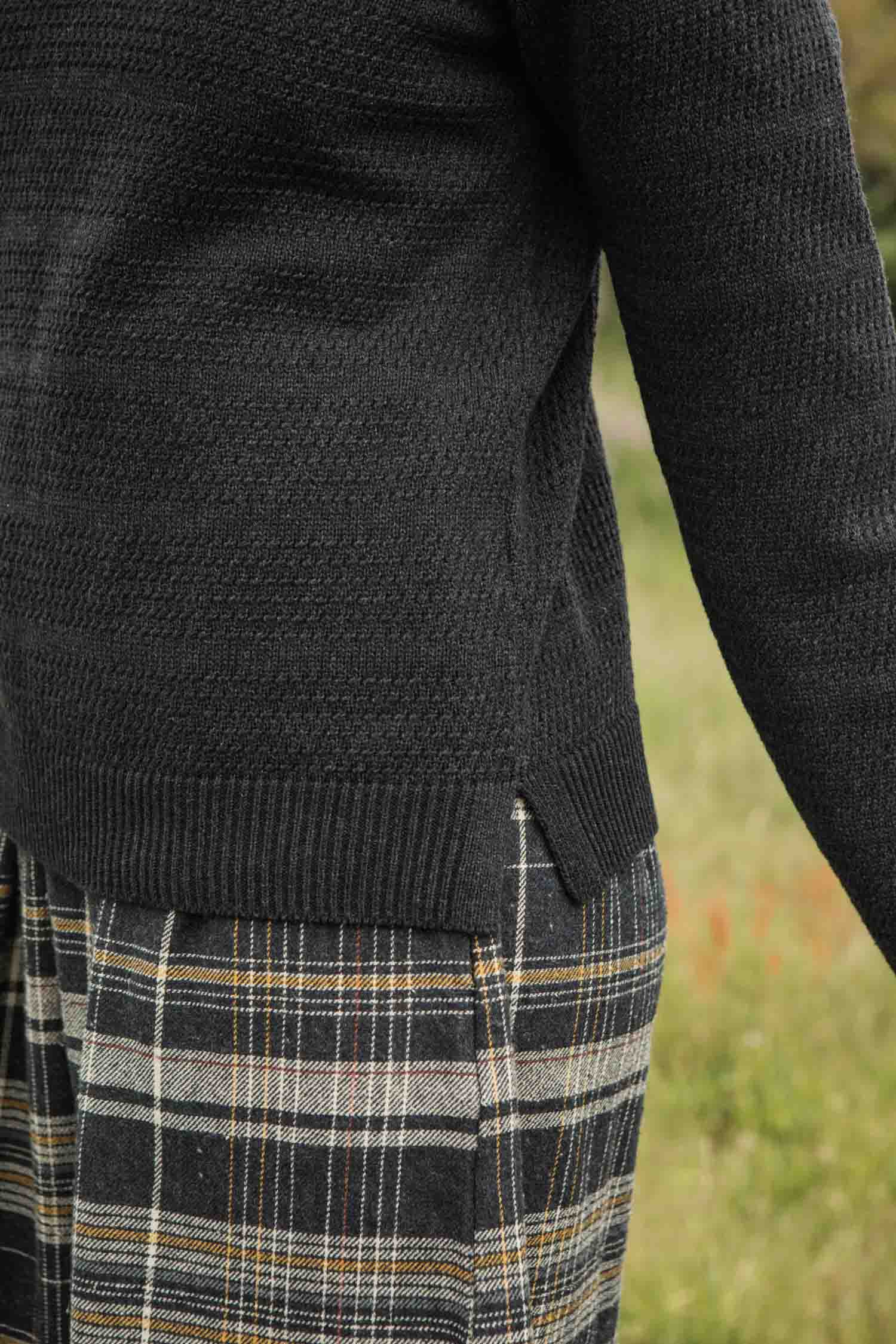 A person stands outside on grass, their side and arm partially visible, wearing the Bridge & Burn Ansel Sweater in Midnight paired with plaid pants.