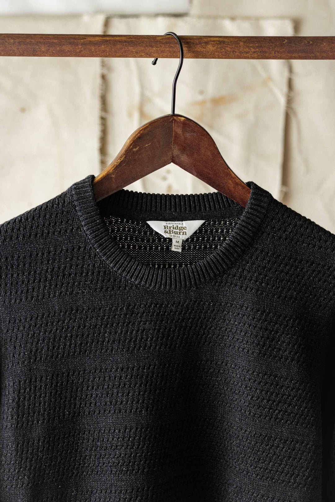 The Ansel Sweater in Midnight by Bridge & Burn, crafted from recycled fibers, hangs on a wooden hanger against a textured background—a sustainable mid-weight essential for any wardrobe.