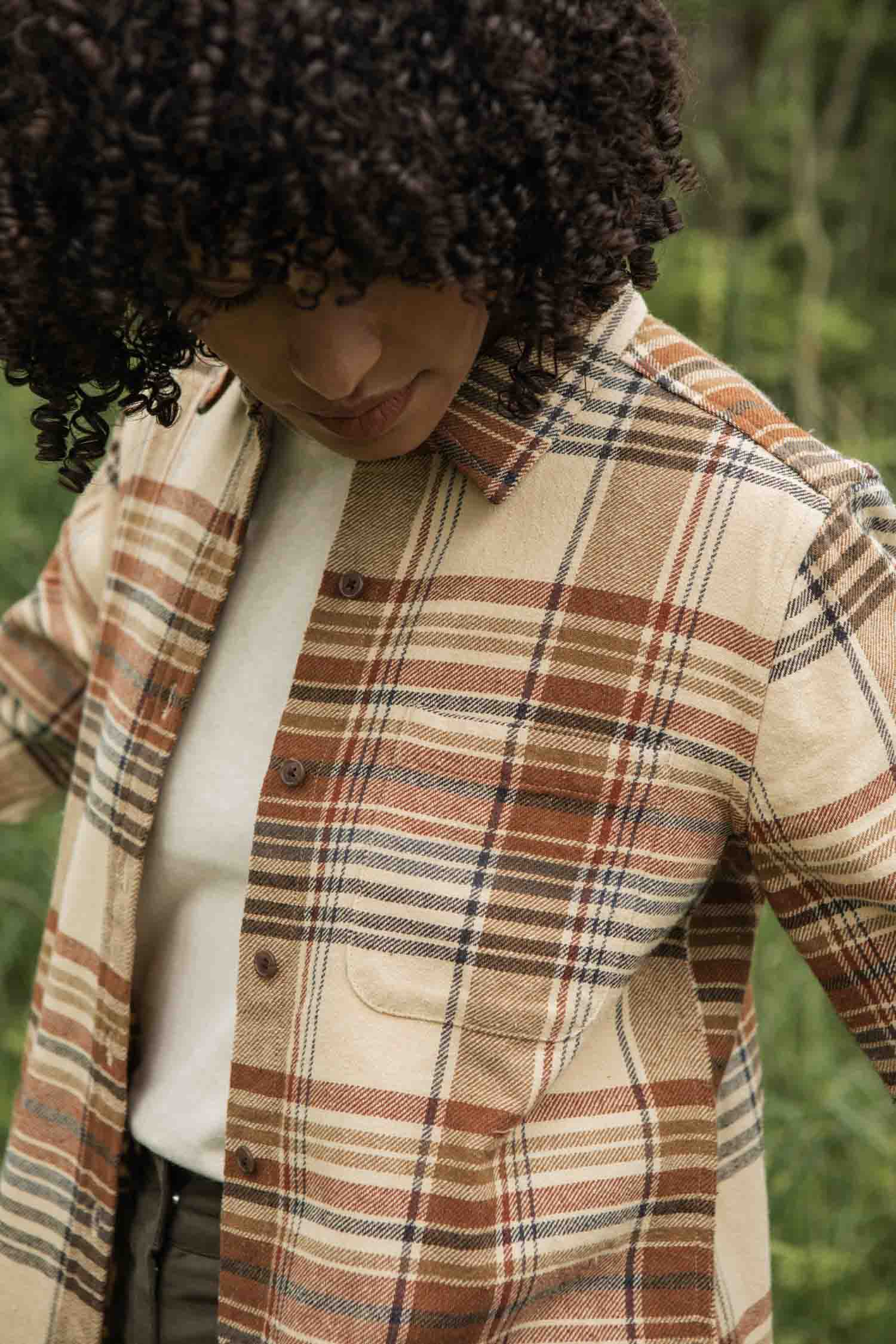 A person with curly hair looks down while wearing the Bridge & Burn Ash Slim Shirt in Latte Plaid over a white t-shirt, standing outdoors with greenery in the background.