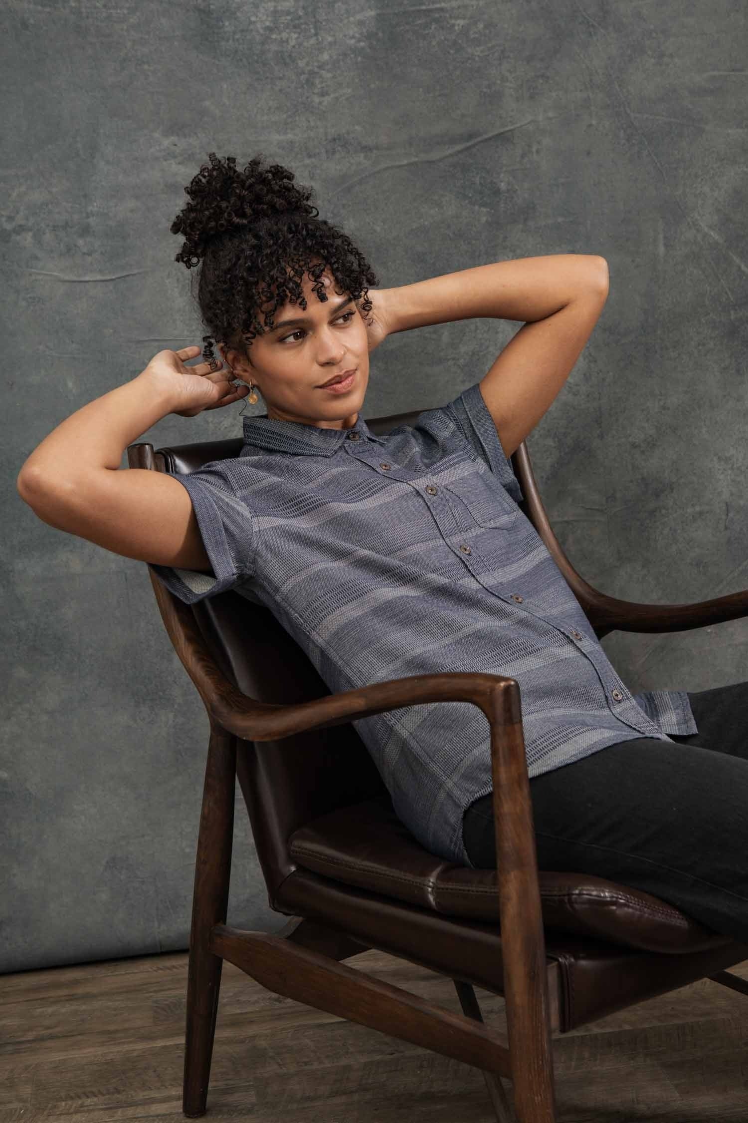 A person with curly hair leans back in a wooden chair, hands behind their head, wearing the Bridge & Burn Bea Shirt / Cascade Weave and dark pants, posed against a textured gray backdrop.