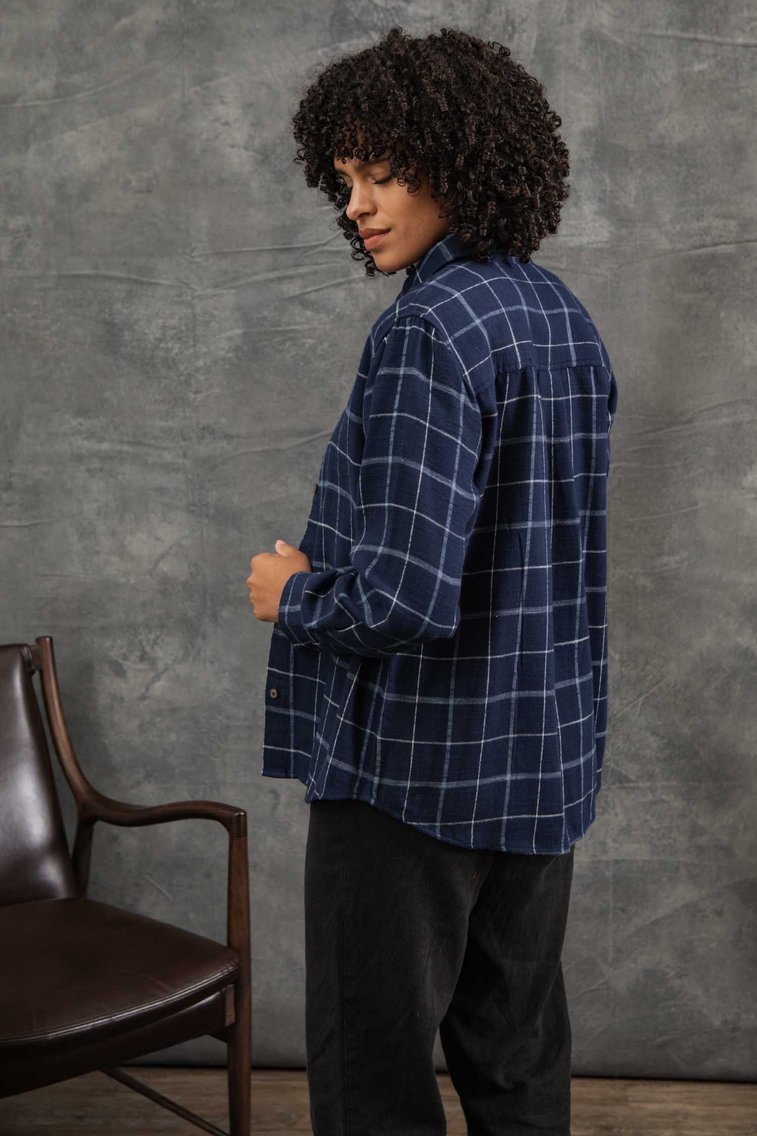 A person with curly hair wears a relaxed fit Bird Shirt / Moonrise Windowpane by Bridge & Burn and black pants, standing sideways and looking over their shoulder. A brown chair sits to the left against a gray textured background.