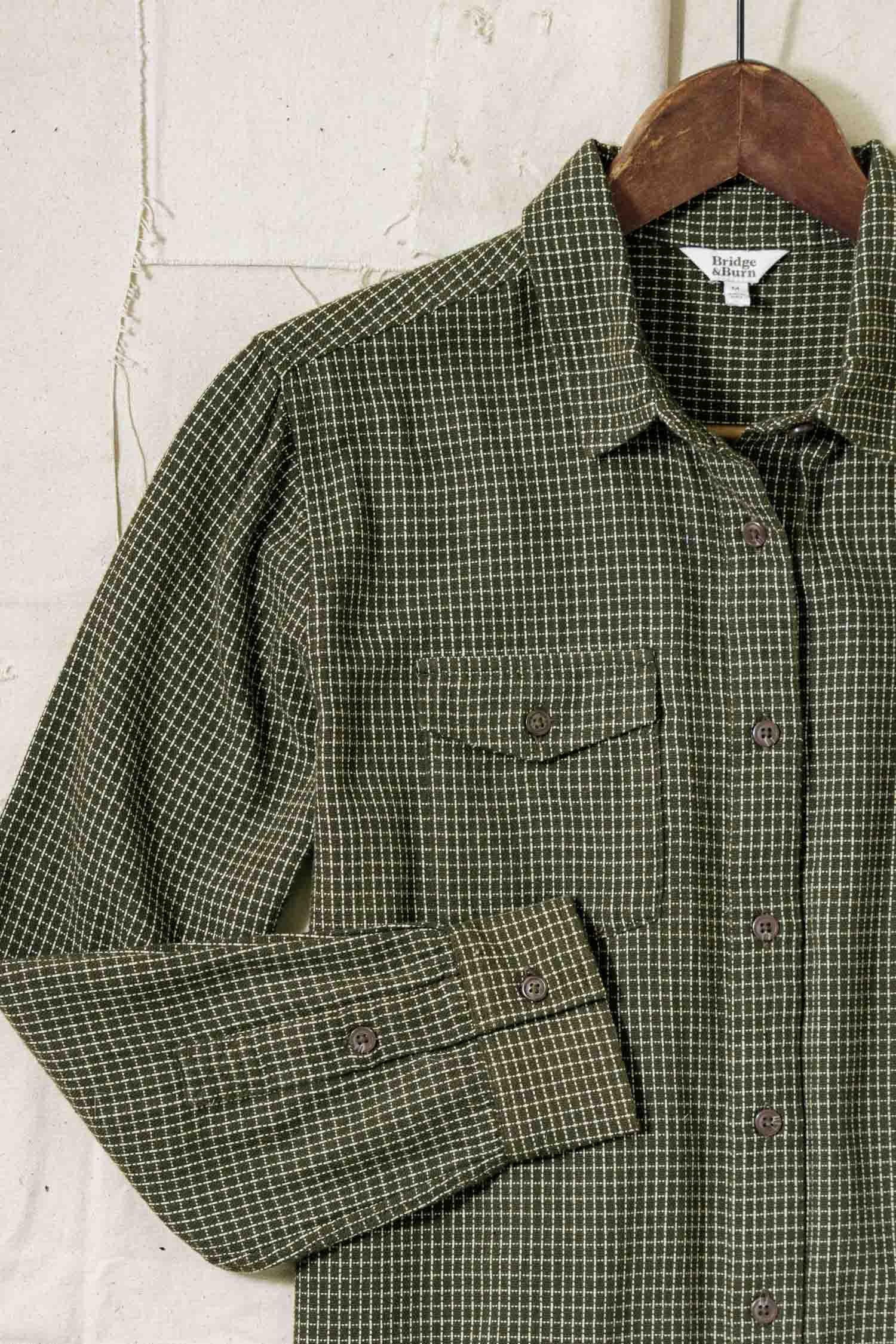 The Bird Shirt / Olive Grid Plaid by Bridge & Burn, a relaxed fit men's shirt with a chest pocket and visible stitching, hangs on a wooden hanger against a light textured wall. The green and white plaid design adds classic style.