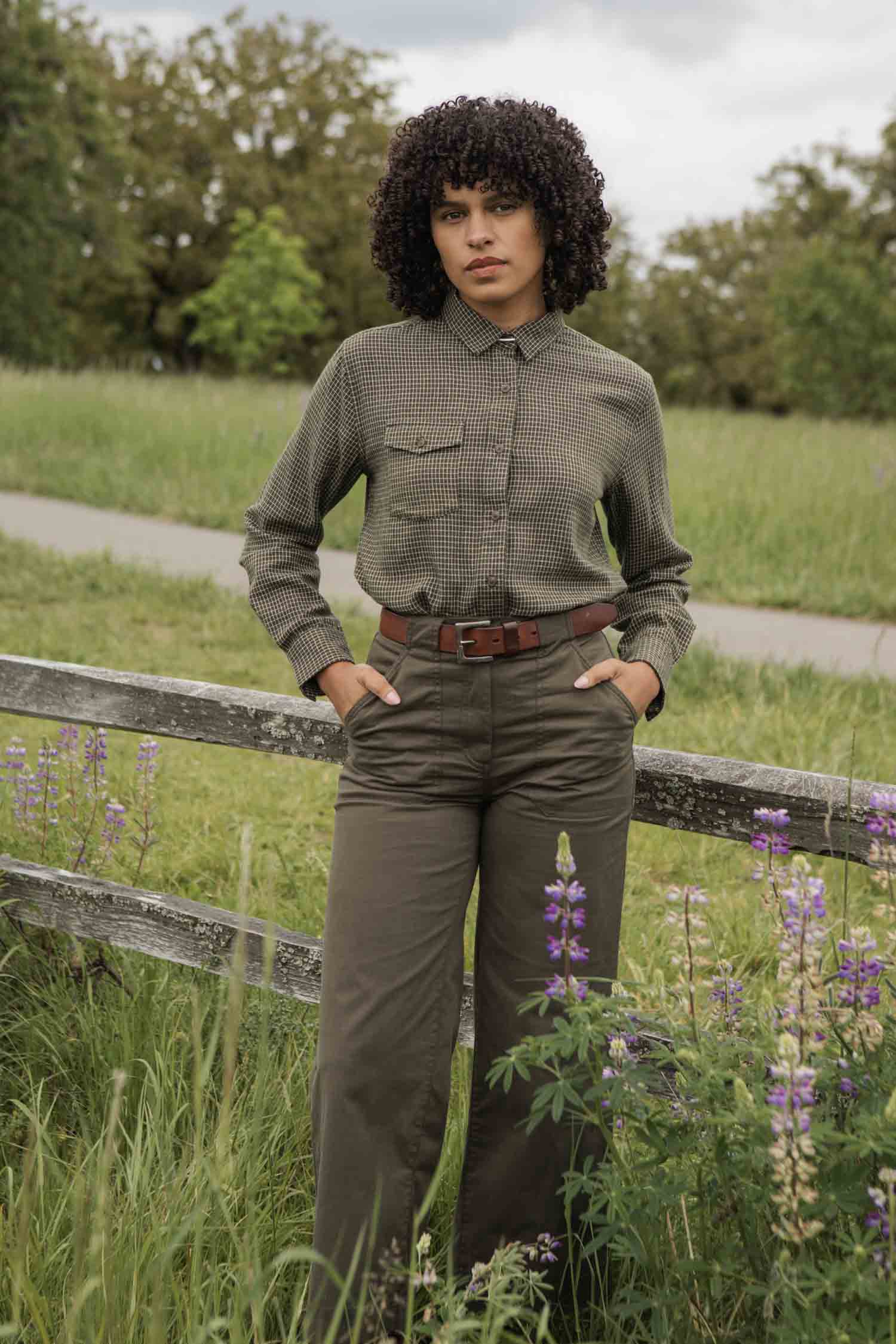 A person with curly hair stands outdoors by a wooden fence, wearing the Bridge & Burn Bird Shirt in Olive Grid Plaid tucked into high-waisted pants with a brown belt. Purple wildflowers, green grass, and trees complete the scenic backdrop.