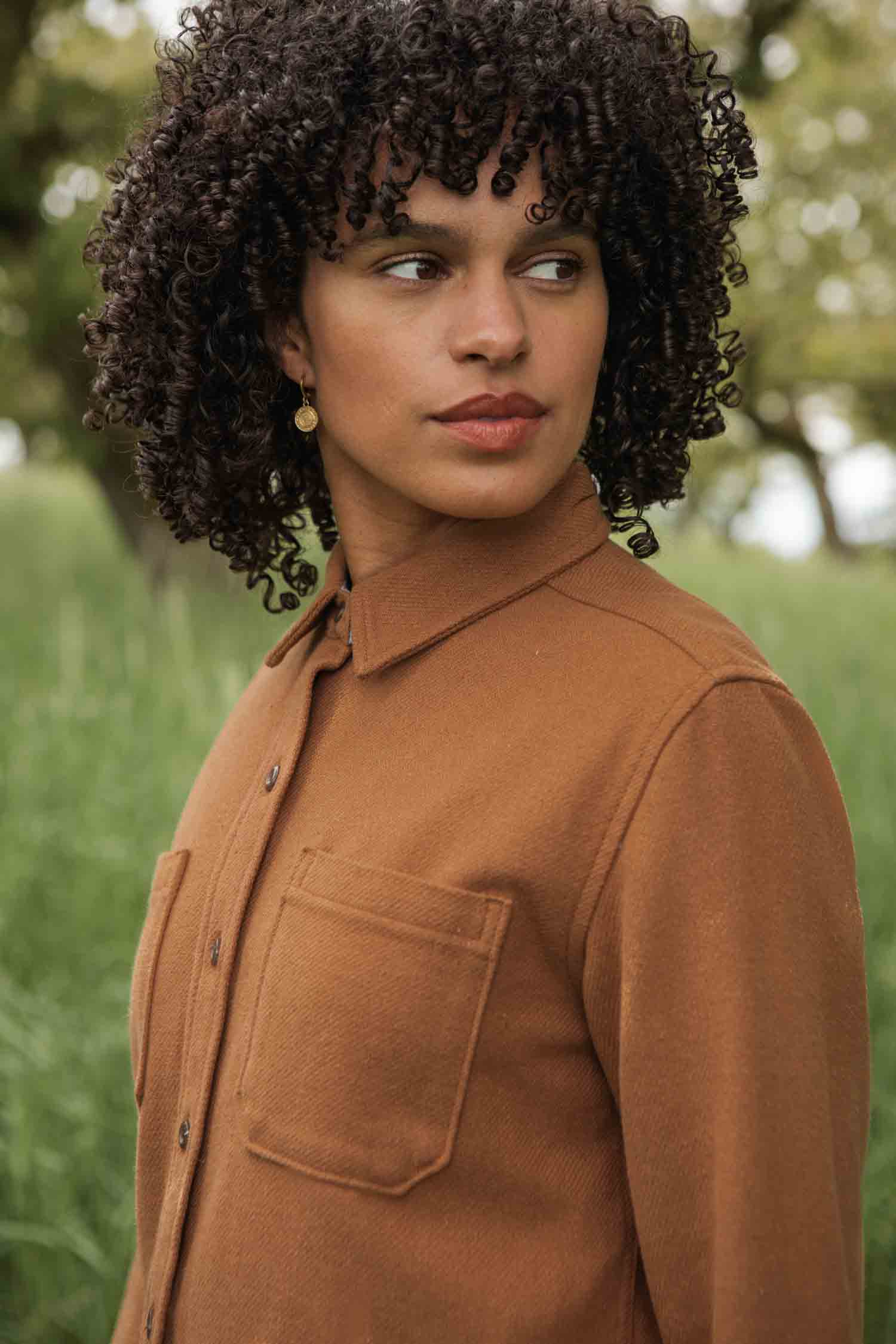 A person with curly dark hair stands outside in green grass and blurred trees, wearing the Bridge & Burn Dawson Overshirt in Camel.