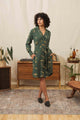 A woman with curly hair wears the Bridge & Burn Emery Shirt Dress in Woodland Bloom and black flats, standing in a stylish living room with mid-century furniture, houseplants, and a decorative rug.