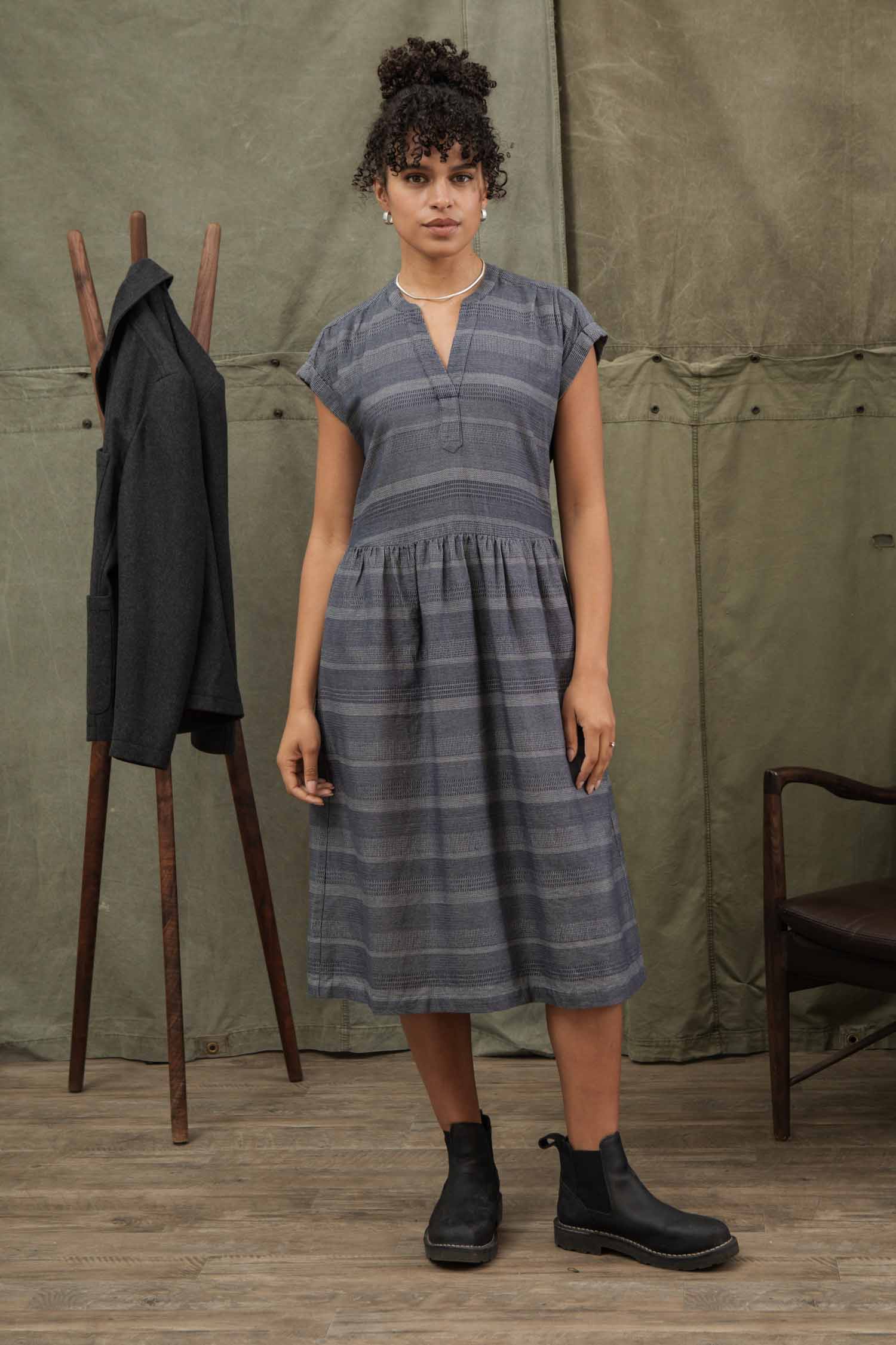 A woman with curly hair in a bun wears the Bridge & Burn Ira Dress in Cascade Weave—a gray, knee-length organic cotton pullover with dolman sleeves—and black ankle boots. Indoors, she stands by a coat rack and nearby brown chair on a wooden floor.