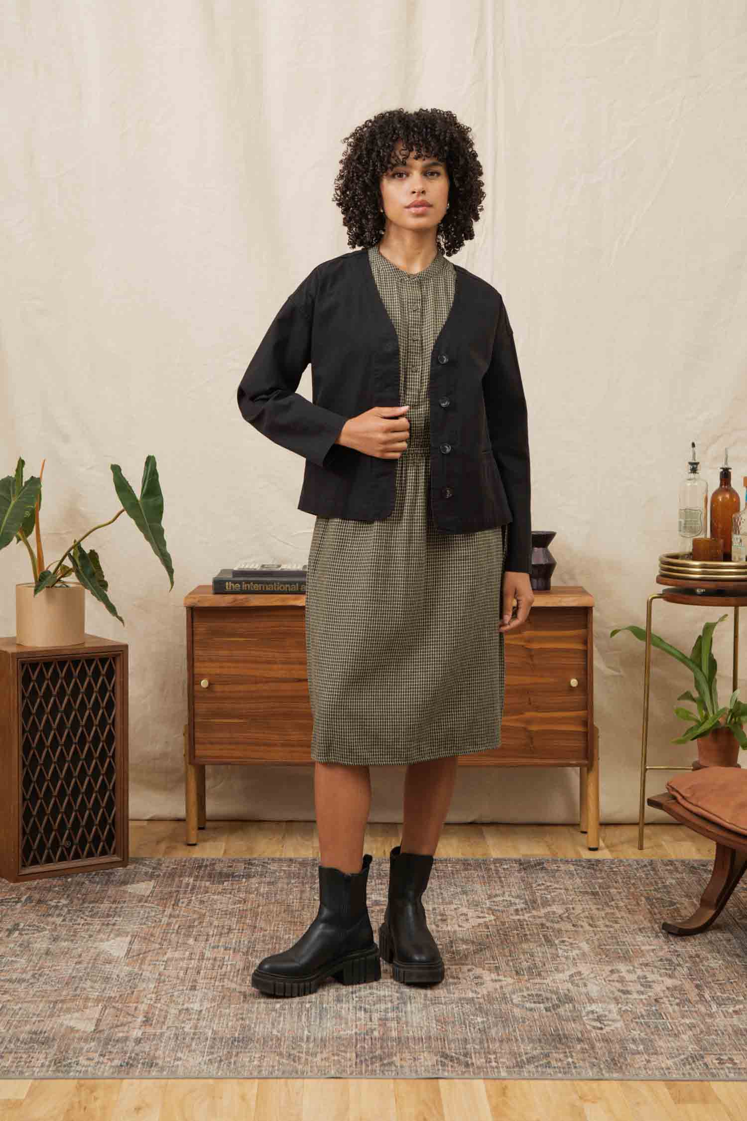 A woman with curly hair stands indoors on a rug, wearing a green plaid dress, black boots, and the Bridge & Burn Lee Jacket in Black Canvas. She is in front of a mid-century console table adorned with plants and bottles.