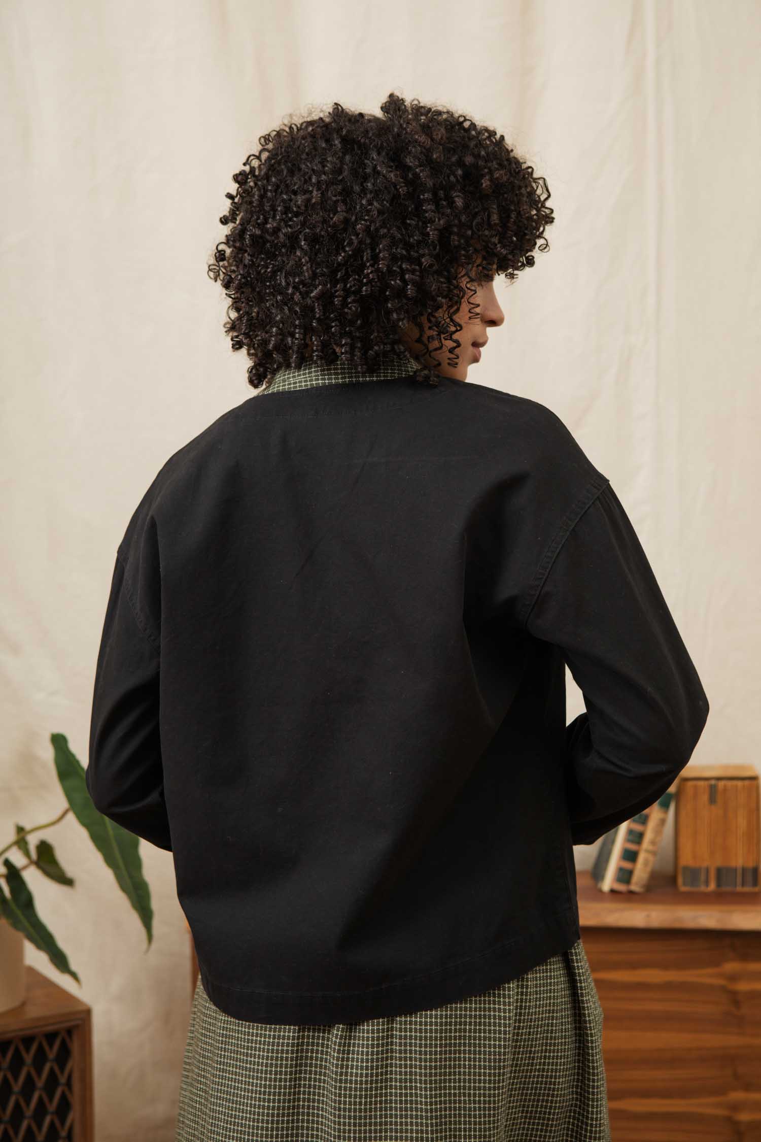 A person with curly hair wears the Bridge & Burn Lee Jacket in Black Canvas over a green checkered shirt, standing with their back to the camera in a room with neutral decor and a vintage radio on a wooden table.
