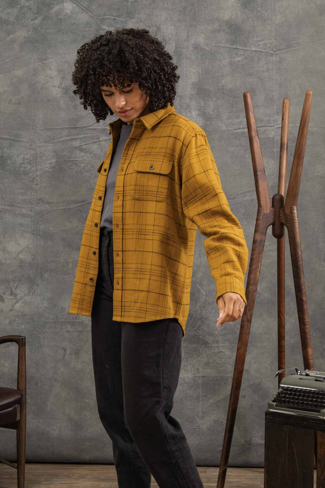 A woman with curly hair wears the Bridge & Burn Lenon Shirt in Golden Horizon Plaid, layered over a gray top and black pants. She stands indoors by a wooden coat rack and typewriter against a gray textured backdrop.