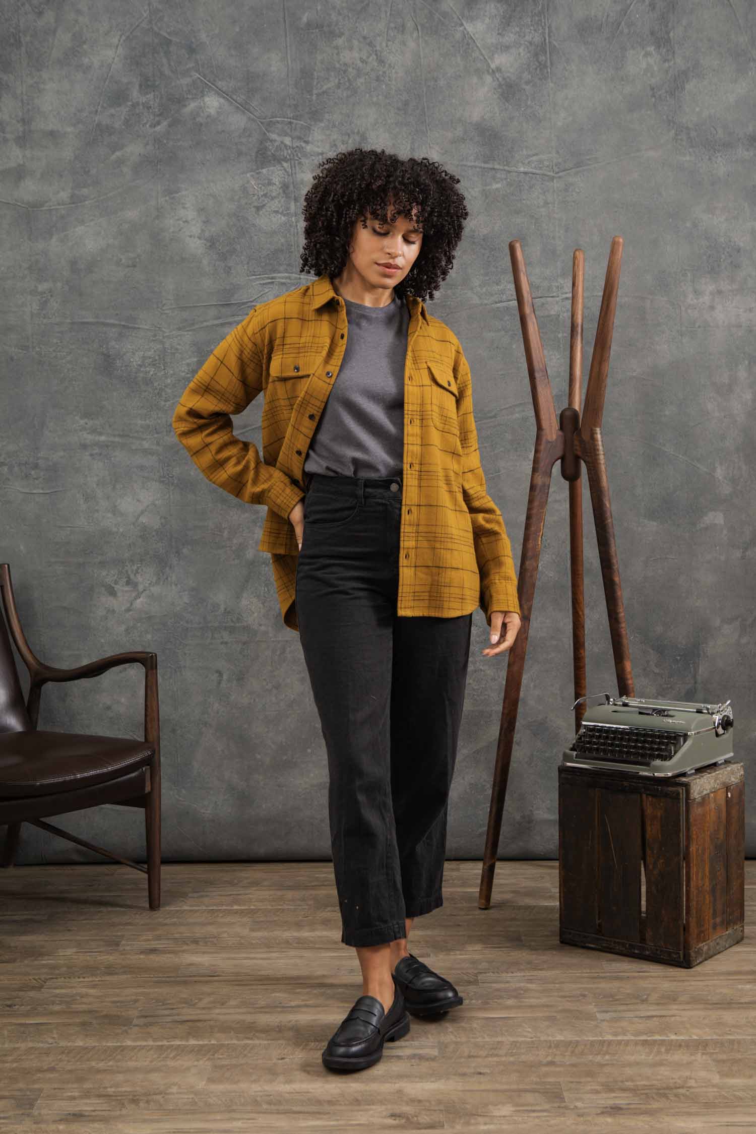 A woman with curly hair wears the Bridge & Burn Lenon Shirt in Golden Horizon Plaid over a gray t-shirt, black pants, and black loafers. She stands indoors near a wooden chair, vintage typewriter, and coat rack.