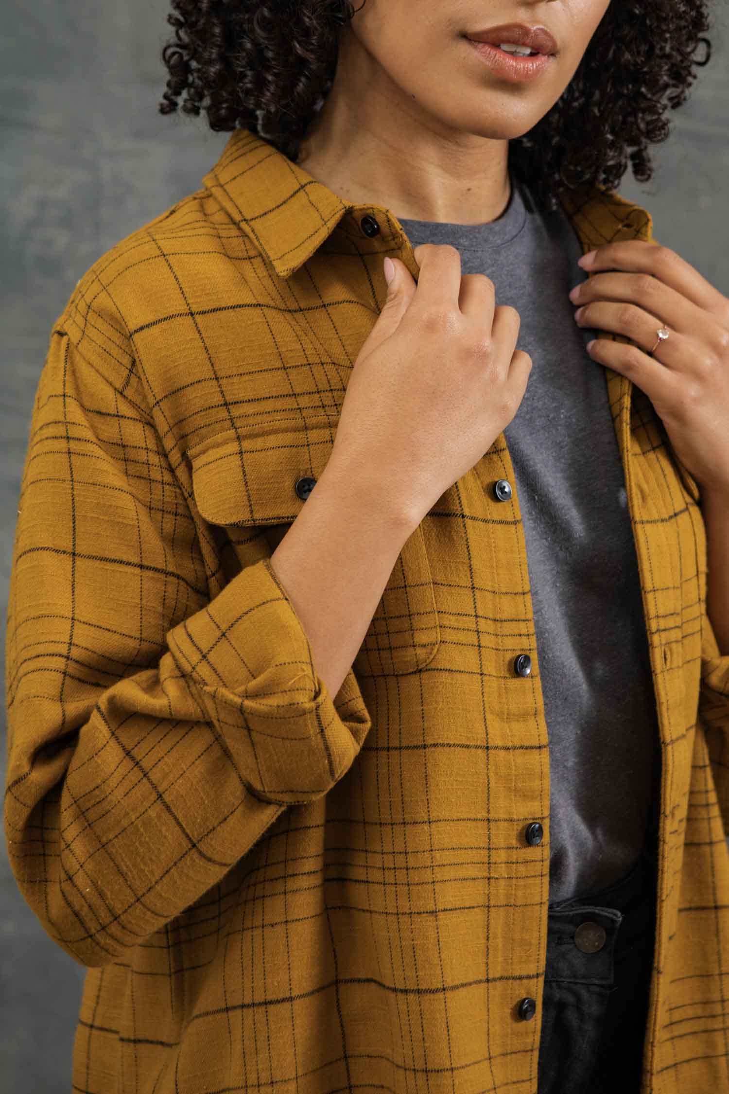 A person with curly hair adjusts the collar of the Bridge & Burn Lenon Shirt in Golden Horizon Plaid, a relaxed fit over-shirt made from organic cotton, worn over a gray t-shirt. Their face is partially visible.