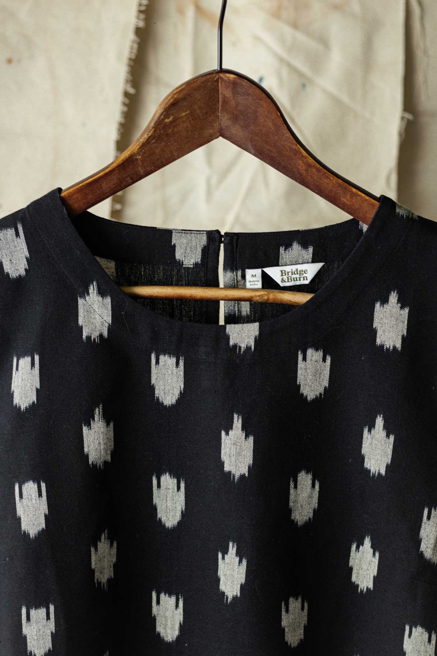 The Leona Shirt / Eclipse Ikat by Bridge & Burn hangs on a wooden hanger against a beige textured fabric background. This boxy blouse features a white abstract dot pattern, and the label reads M/L and Bridge & Burn.