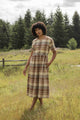 A person wears the Bridge & Burn Perry Dress in Latte Plaid, a short-sleeved, midi-length style with a gathered skirt, standing barefoot in a grassy field with trees and a wooden fence under a cloudy sky.