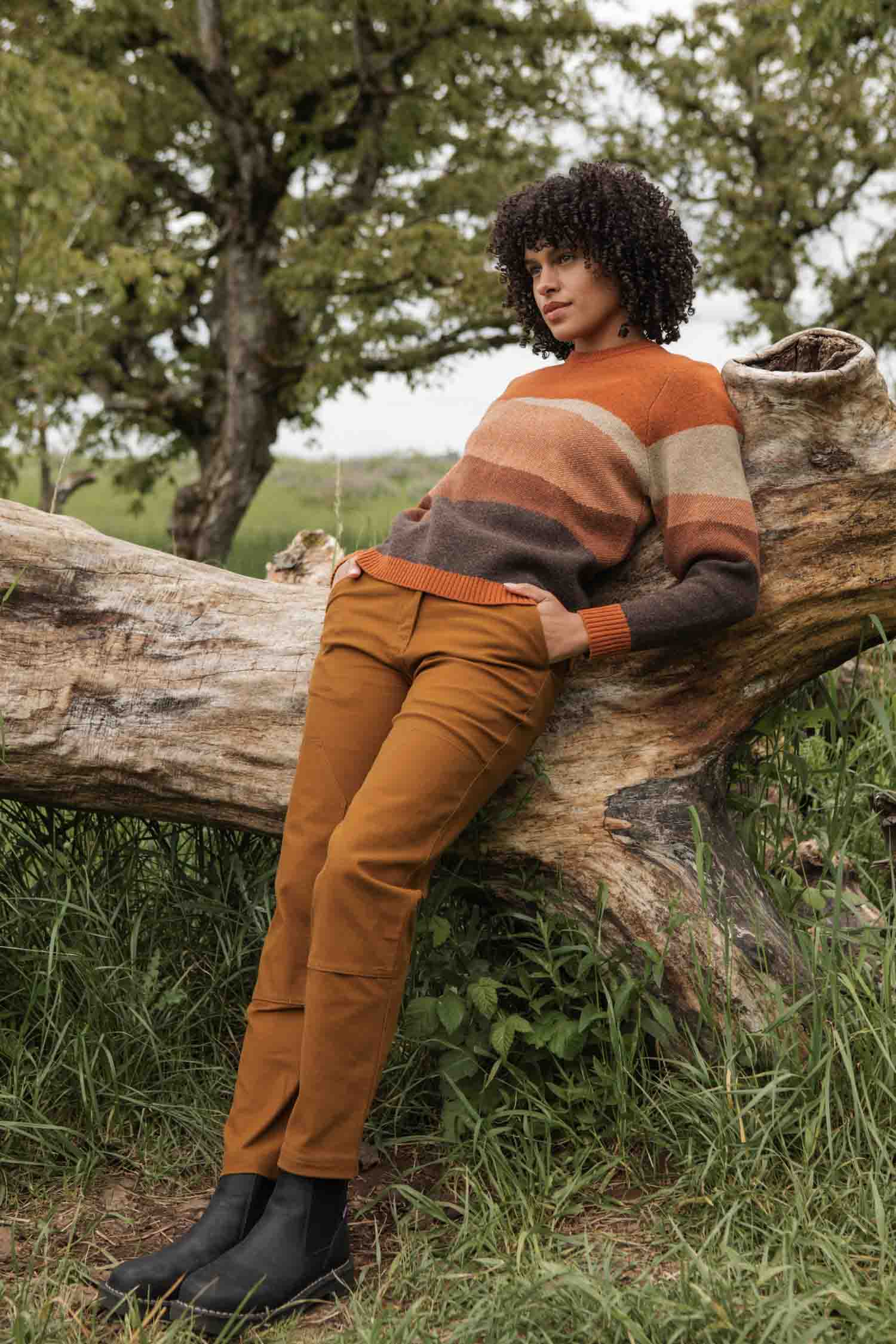 A person with curly hair leans against a fallen tree outdoors, wearing the Bridge & Burn Petra Sweater in Sandstone, rust-colored pants, and black boots, surrounded by grass and trees.