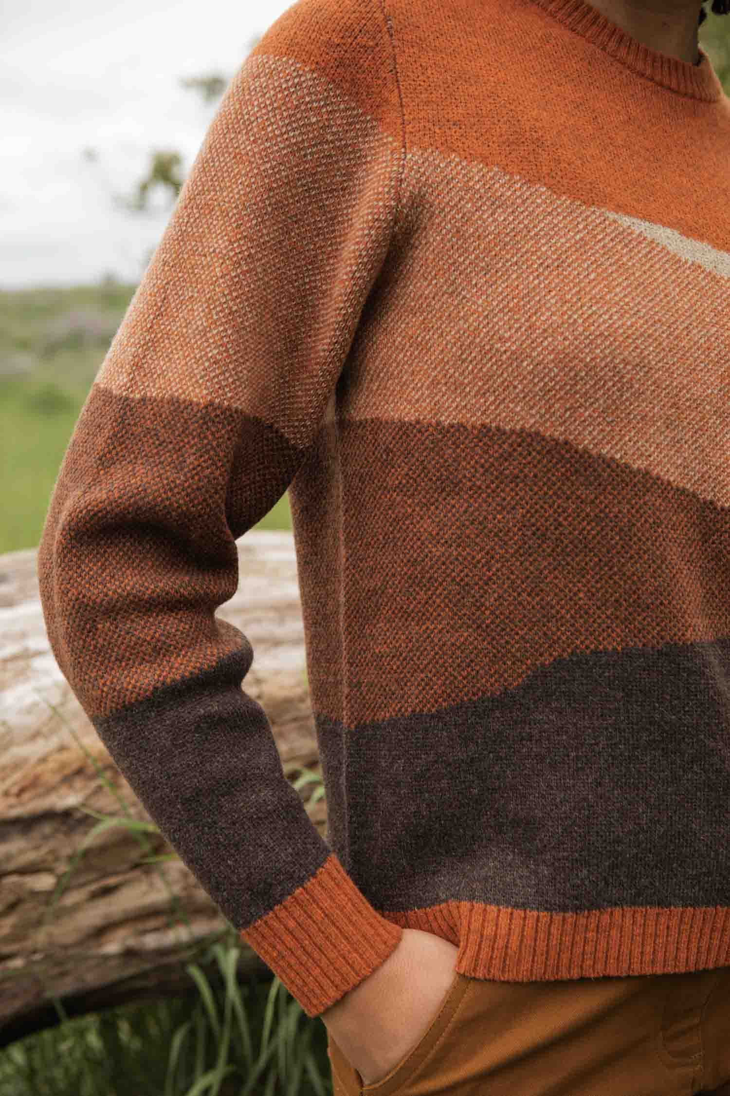 A person wears the Bridge & Burn Petra Sweater in Sandstone—featuring warm earthy stripes of orange and brown—standing outdoors with one hand in their pocket. A blurred fallen log and greenery appear in the background.