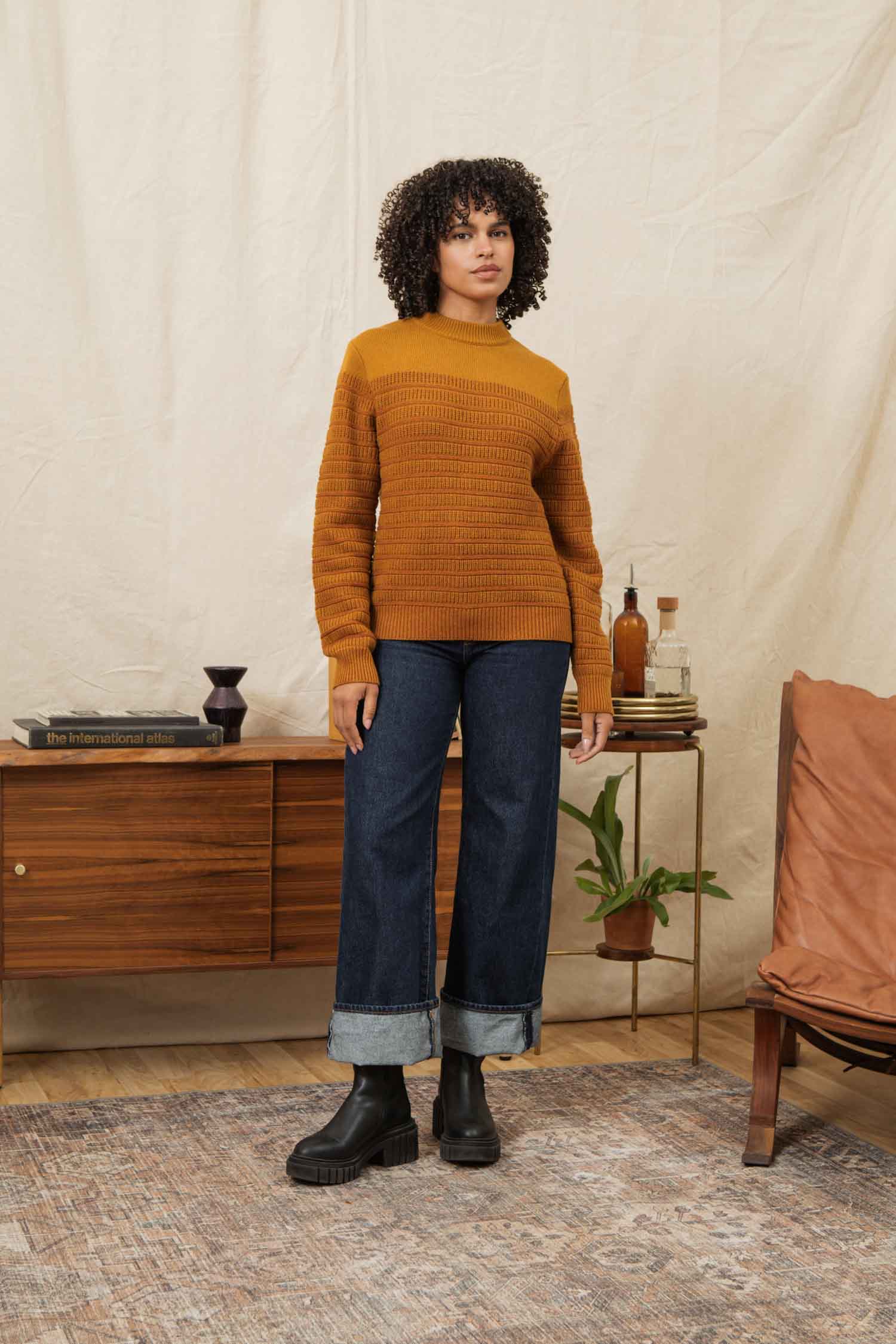 A woman with curly hair models the Bridge & Burn Sabine Sweater in Goldenrod Stripe, paired with wide cuffed jeans and black boots. She stands indoors by a wooden sideboard with books, decor, and a potted plant in a cozy, neutral-toned setting.