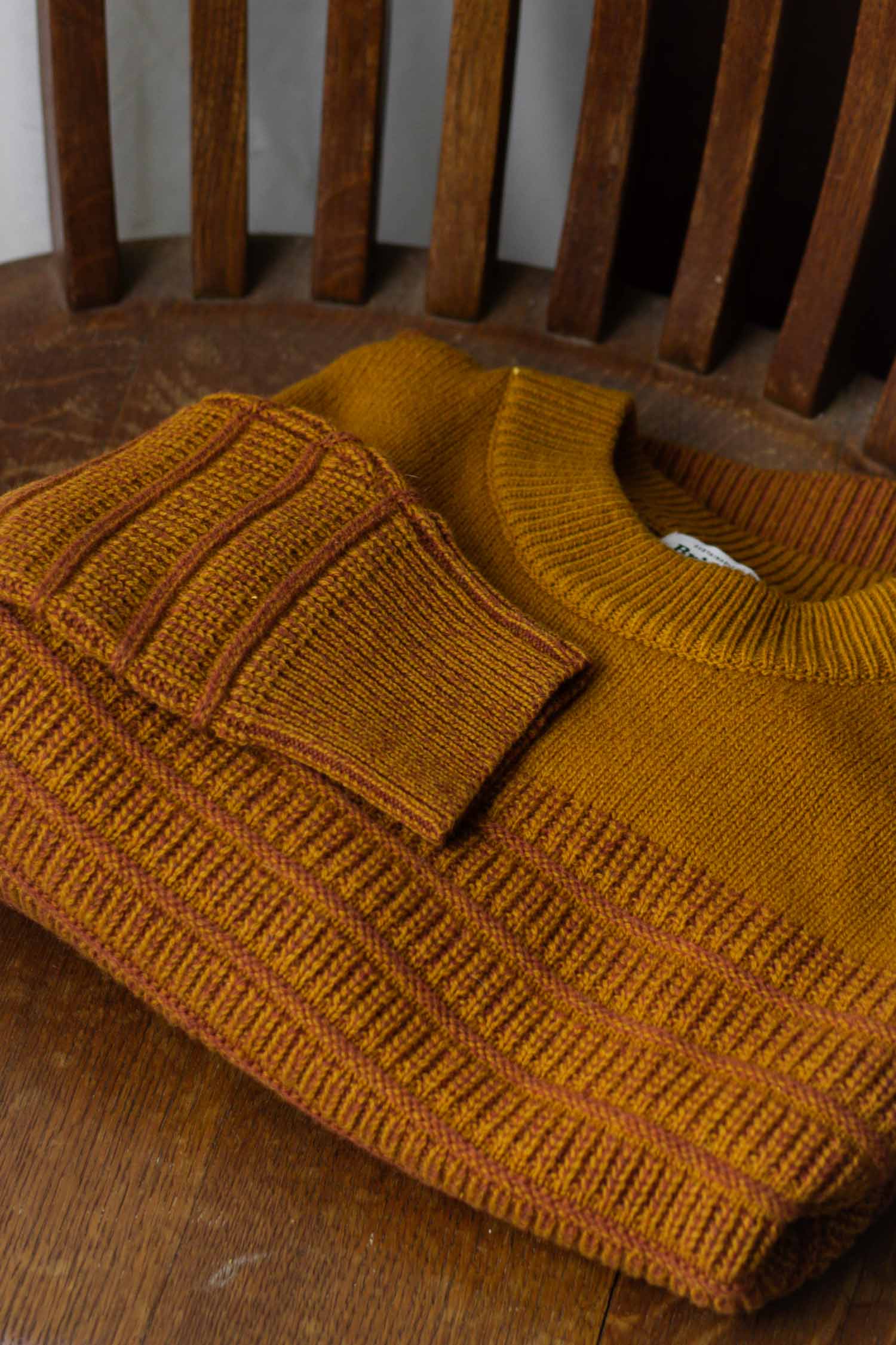 The Bridge & Burn Sabine Sweater in Goldenrod Stripe, a heavyweight knit with ribbed cuffs and hem, is folded neatly on a wooden chair with vertical slats, placed atop a wooden surface.